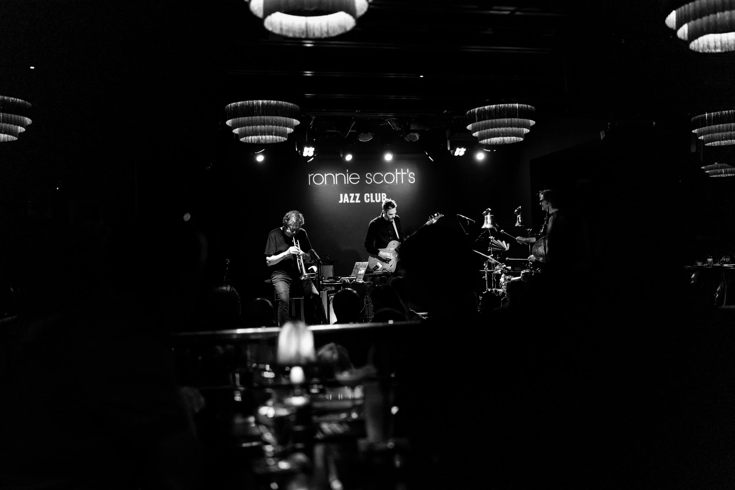 Black and white photo of a jazz band performing on stage at Ronnie Scott's Jazz Club, with three musicians playing instruments under stage lights and the club's name displayed on the background screen.