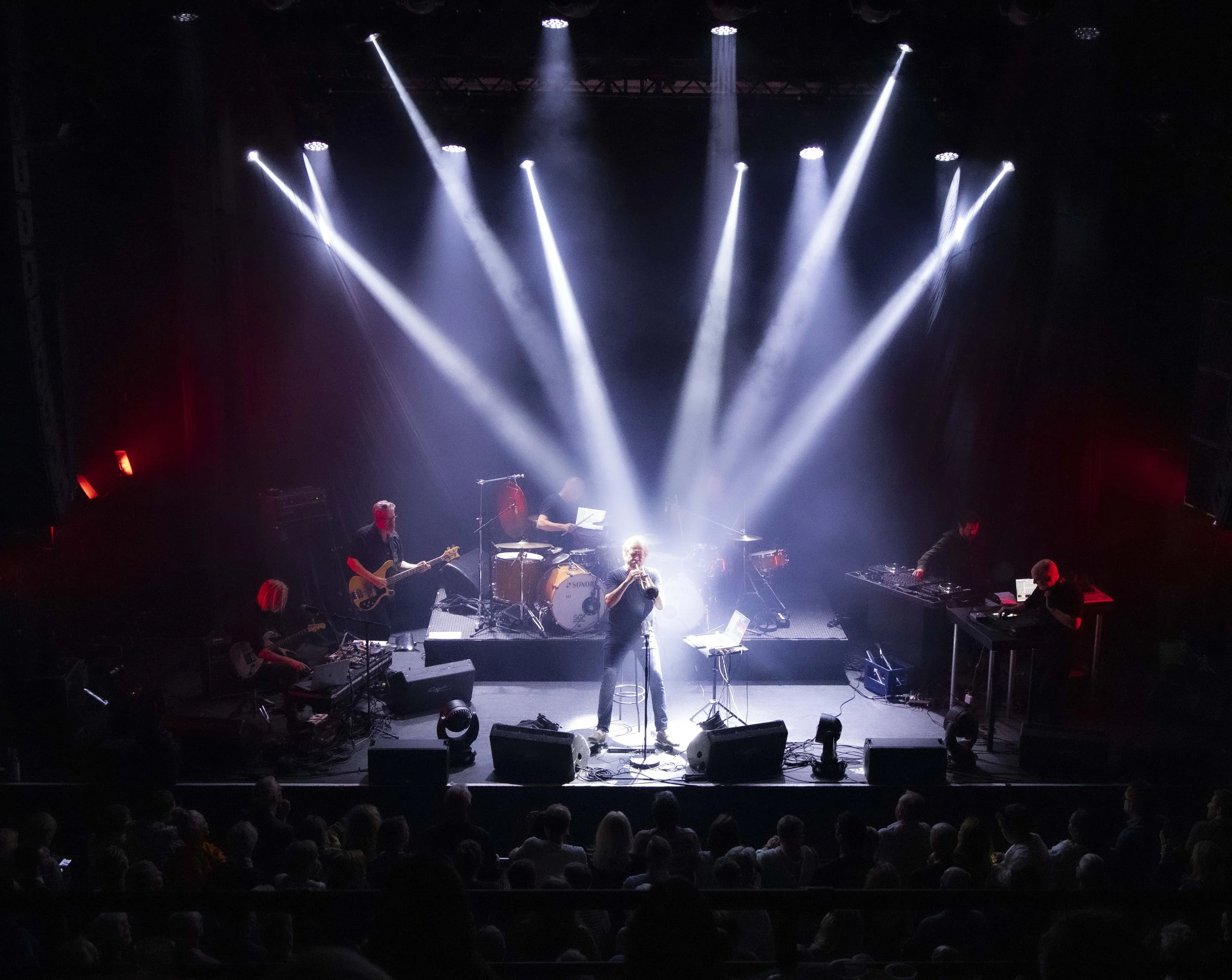 Nils Petter Molvaer 's Khmer performing on stage with bright white lights shining down on them, with the audience in foreground.