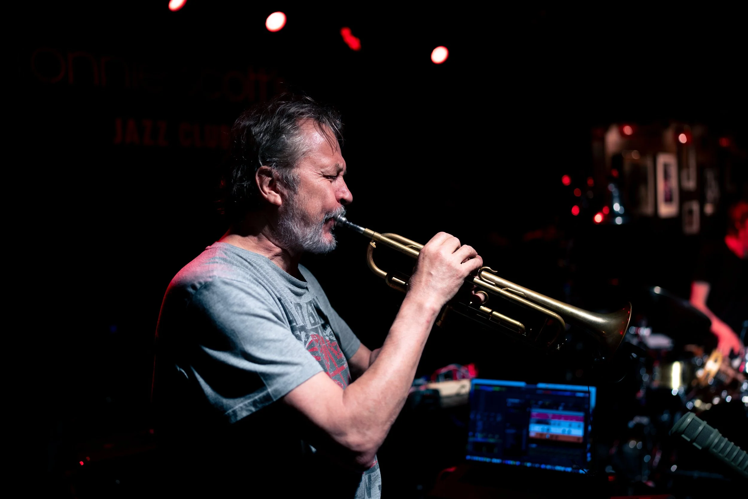 Nils Petter Molvaer playing trumpet on stage at jazz club with dark background and dim red lighting.