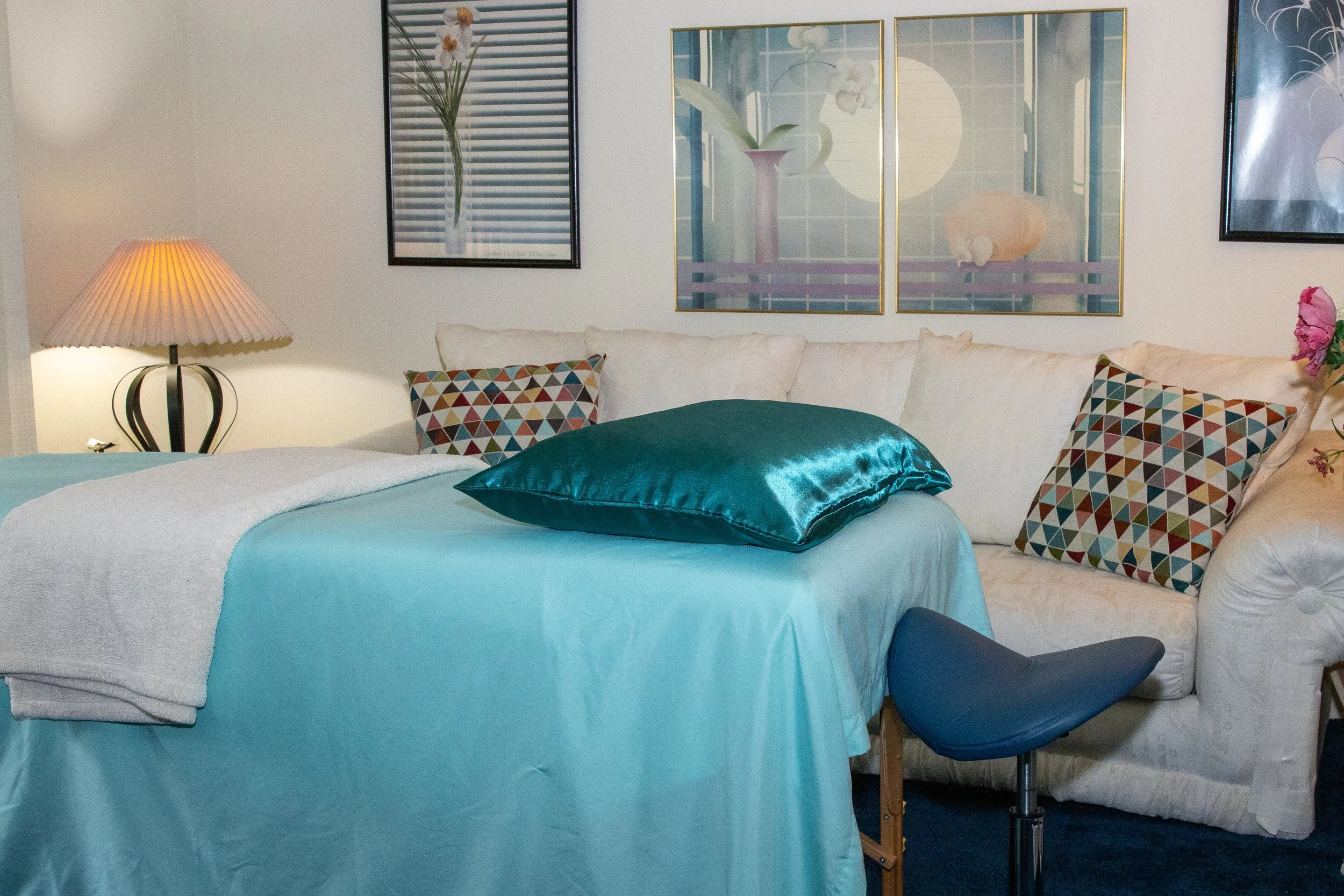 A cozy bodywork suite featuring a massage table with teal satin sheets and pillow, a cushioned stool, soft lighting, a cream sofa with patterned pillows, and calming floral artwork on the walls.
