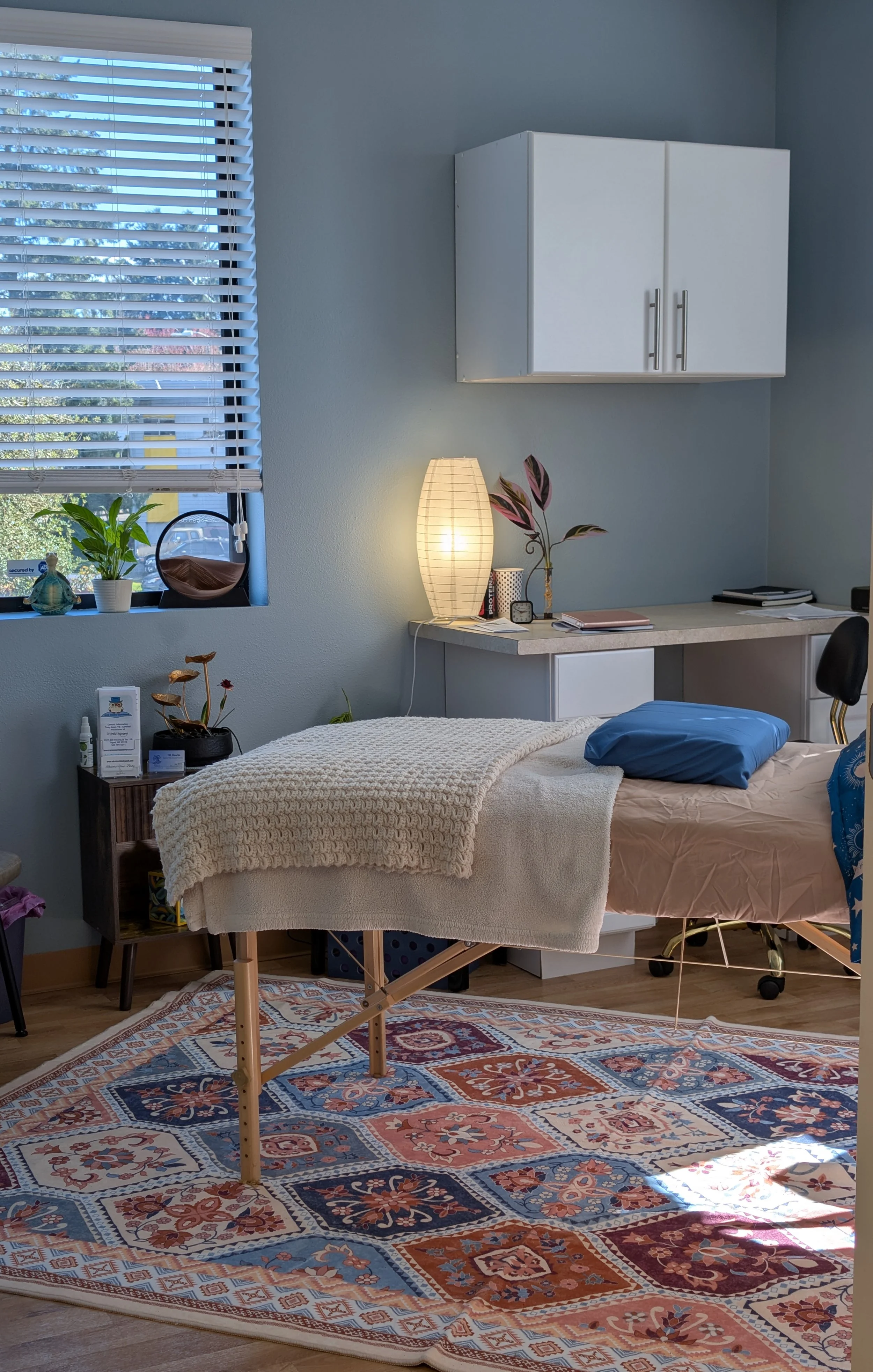 A room with a massage table covered with a white blanket, a blue pillow, and a colorful patterned area rug on the floor. There is a window with blinds, a small side table with plants, and a desk with decorative items, a lamp, and a chair.
