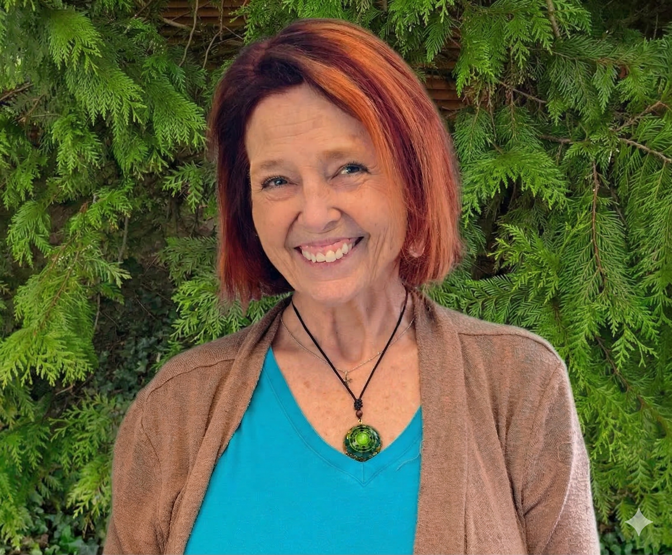 Smiling woman with red hair wearing a blue shirt, brown cardigan, and a green pendant necklace, standing in front of green foliage.