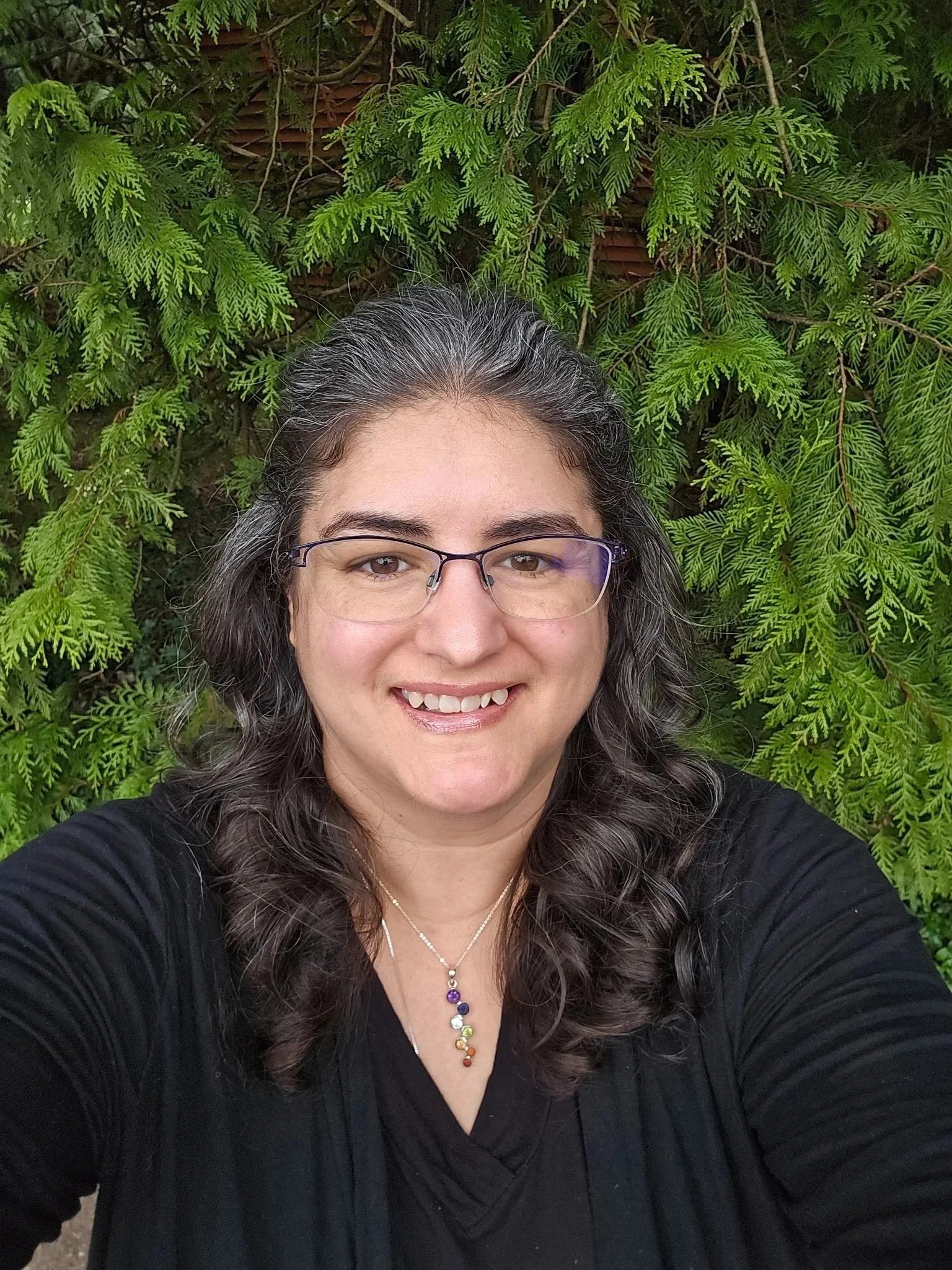 A woman with glasses and long, curly dark hair smiling in front of green foliage.