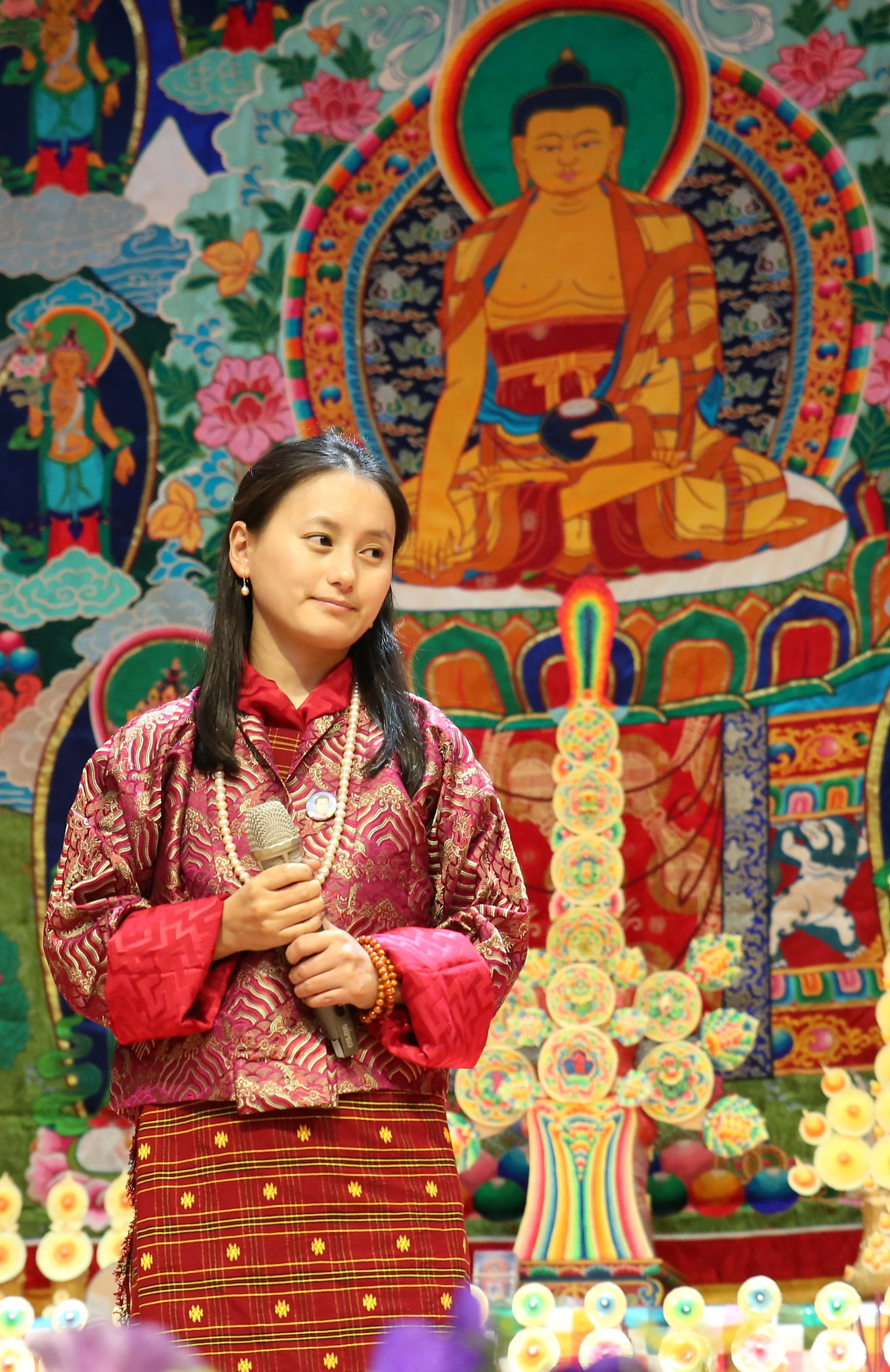 A woman dressed in traditional colorful clothing, holding a microphone, standing in front of an elaborate, vibrant Buddhist or cultural tapestry with images of deities and symbols.