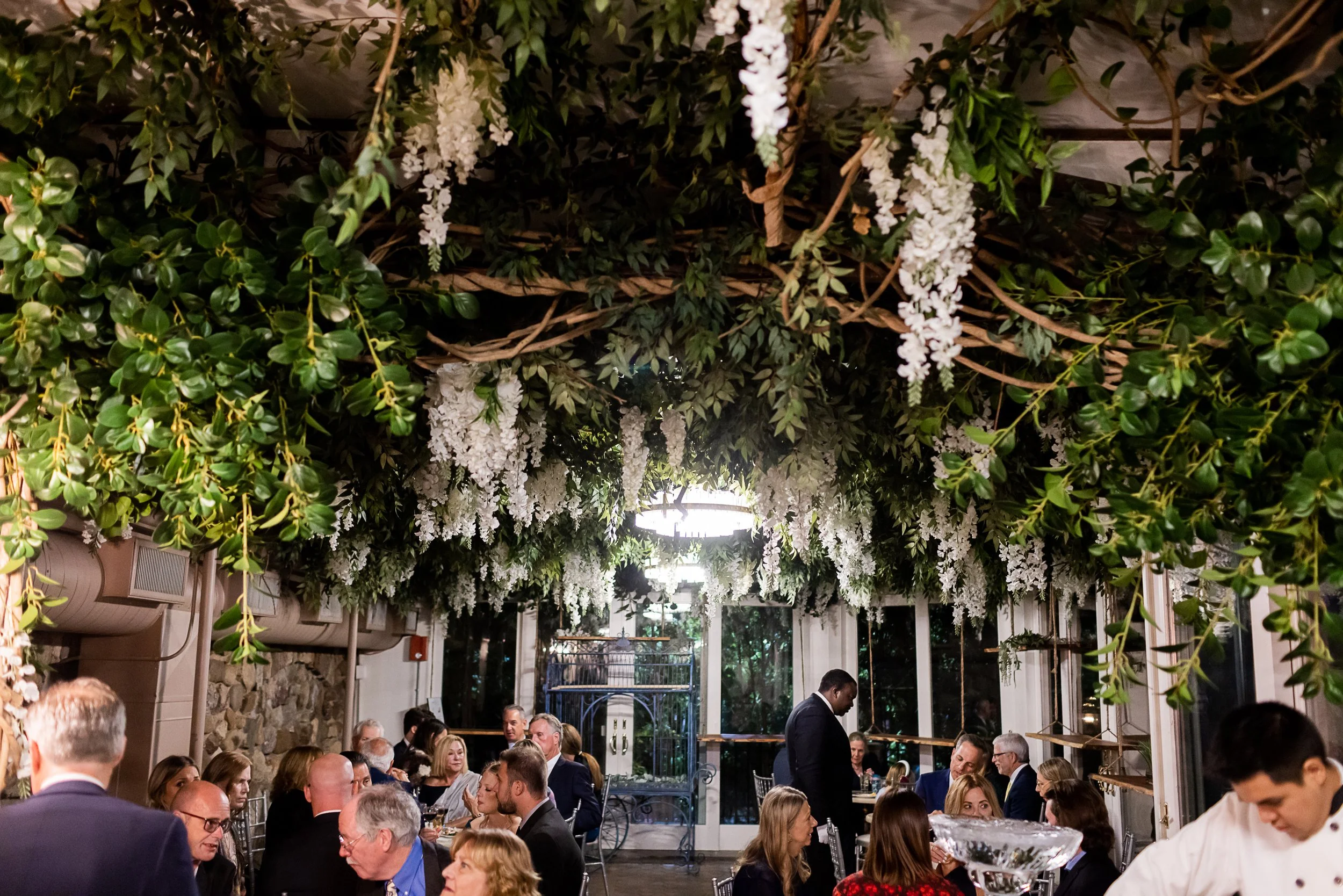 Indoor celebration event with guests dining under a hanging floral and greenery decor ceiling.