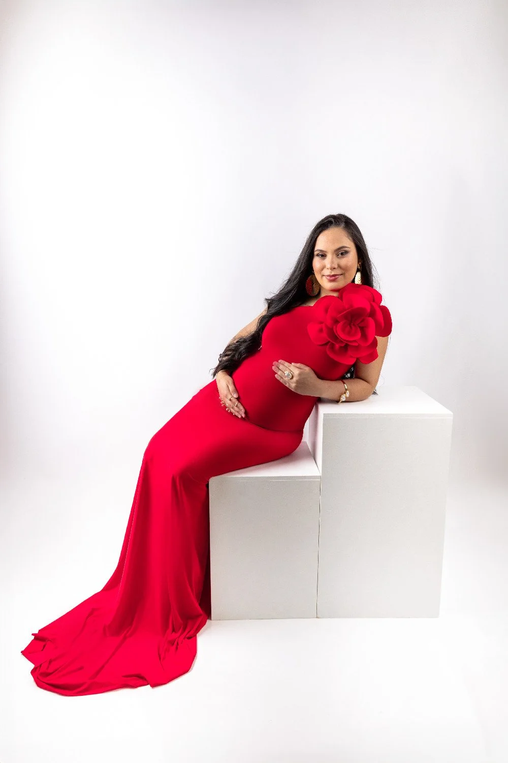 Femme en robe rouge longue avec une grande fleur rouge sur l'épaule, posant avec élégance sur des blocs blancs dans un studio blanc.