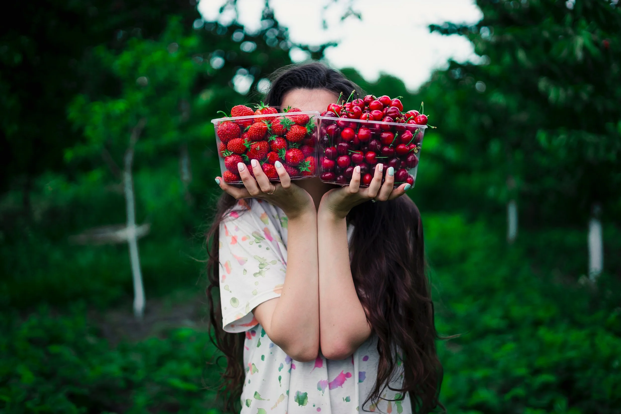 Vrouw met lang bruin haar houdt twee grote glazen met aardbeien en kersen voor haar gezicht, achtergrond van een bos of groen landschap.