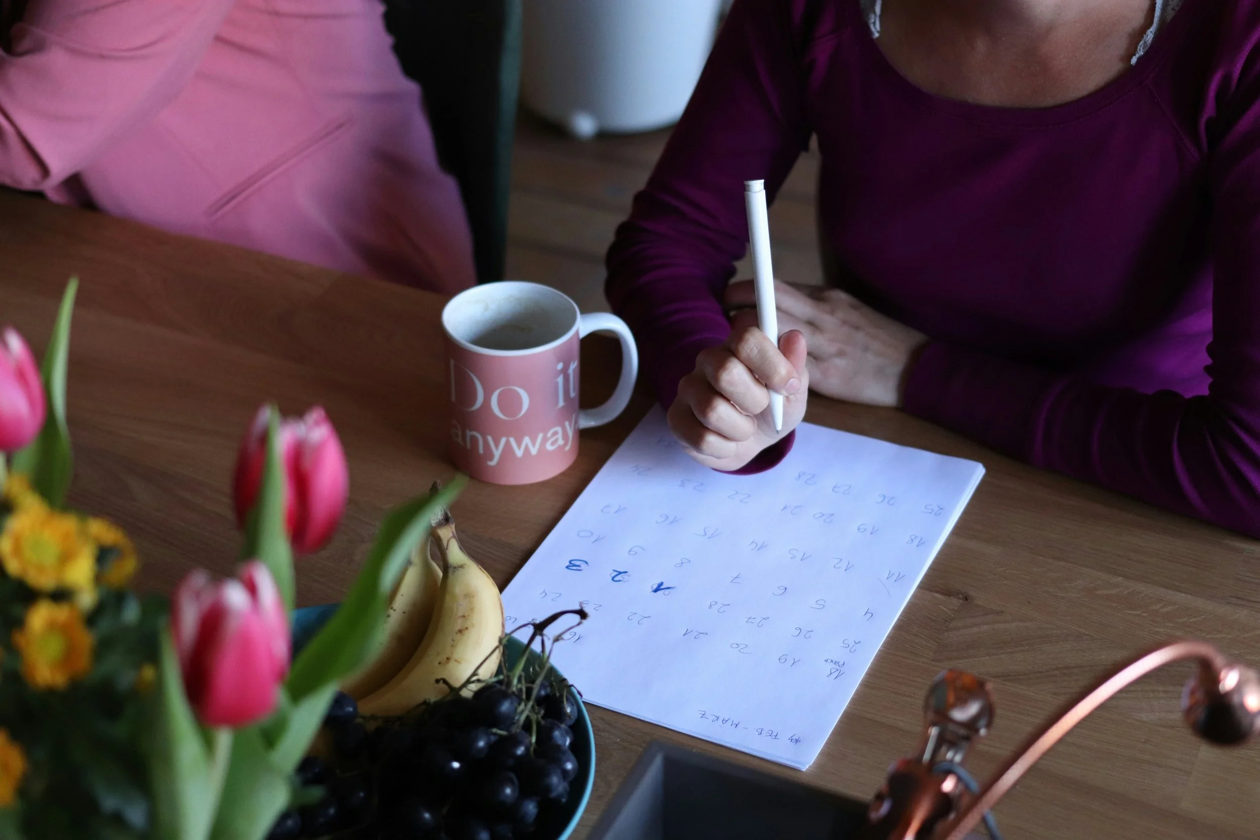 Twee mensen schrijven in een notitieboekje op een houten tafel, met een mok en fruit, waaronder bananen en bessen. In de voorgrond bevinden zich bloemen.