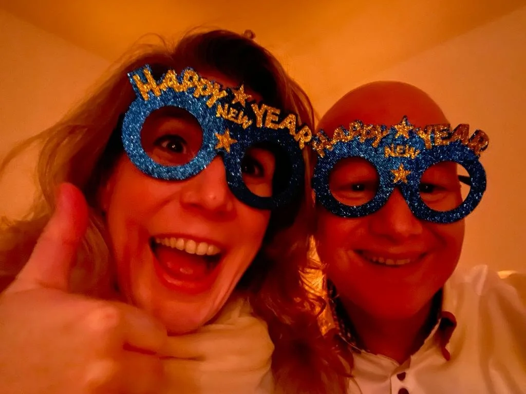 Twee mensen dragen blauwe glitterbrillen met 'HAPPY NEW YEAR' en sterren, vieren nieuwjaar, lachen, indoors met oranje gloed.