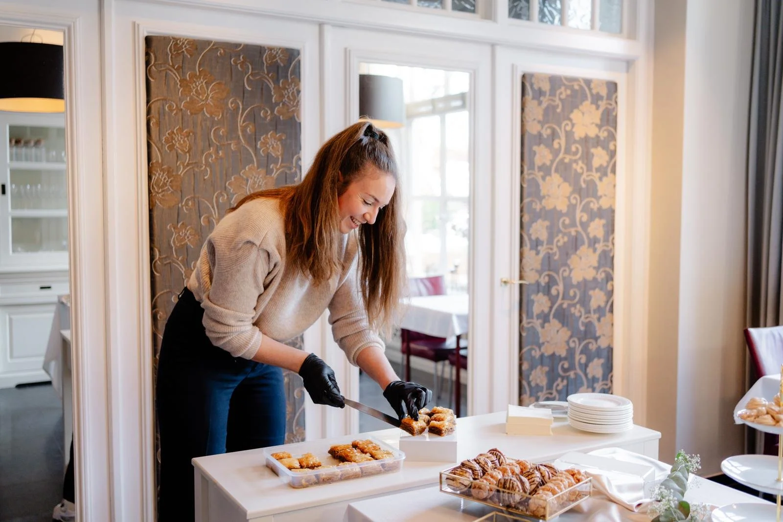 Shivah die haar gebak klaar zet op een witte tafel in een huiselijke eetruimte met bloemendecoratie.