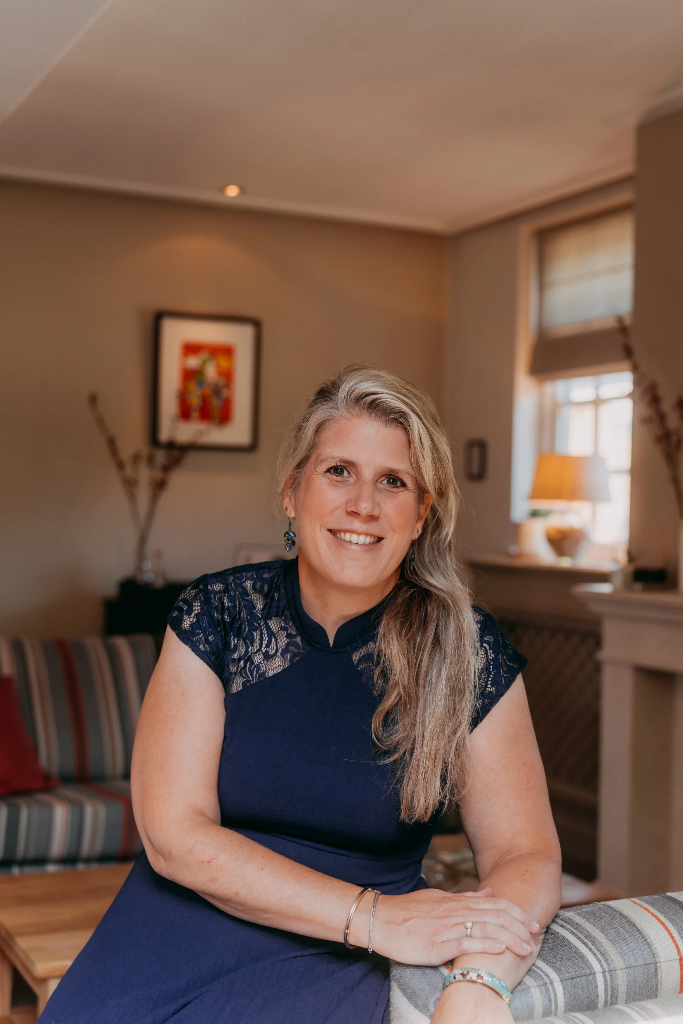 Vrouw met lang blond haar, glimlach, zittend aan een tafel in een woonkamer met afgestemde inrichting