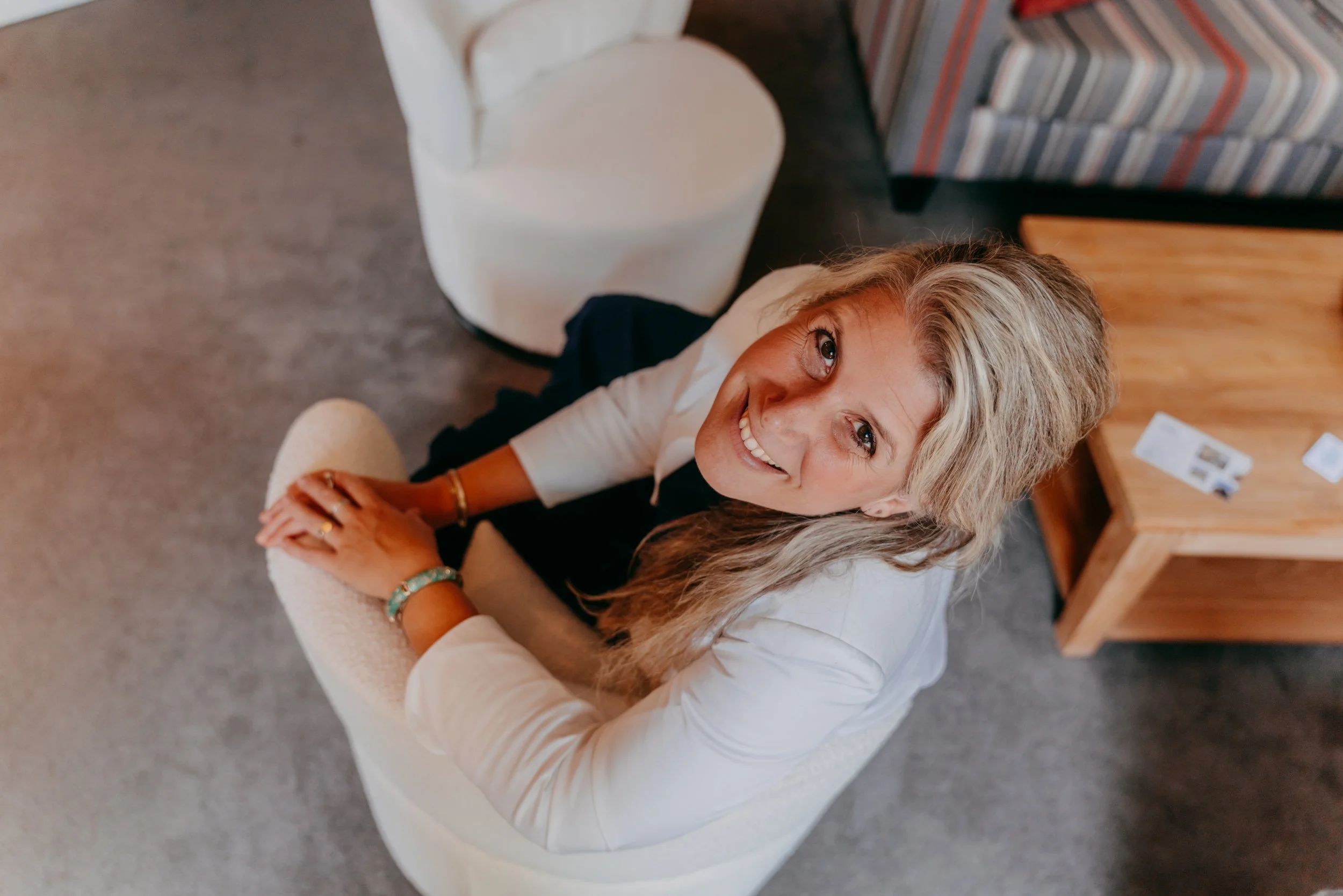 Een lachende vrouw met lang, golvend blond haar zit op een witte stoel en kijkt omhoog.