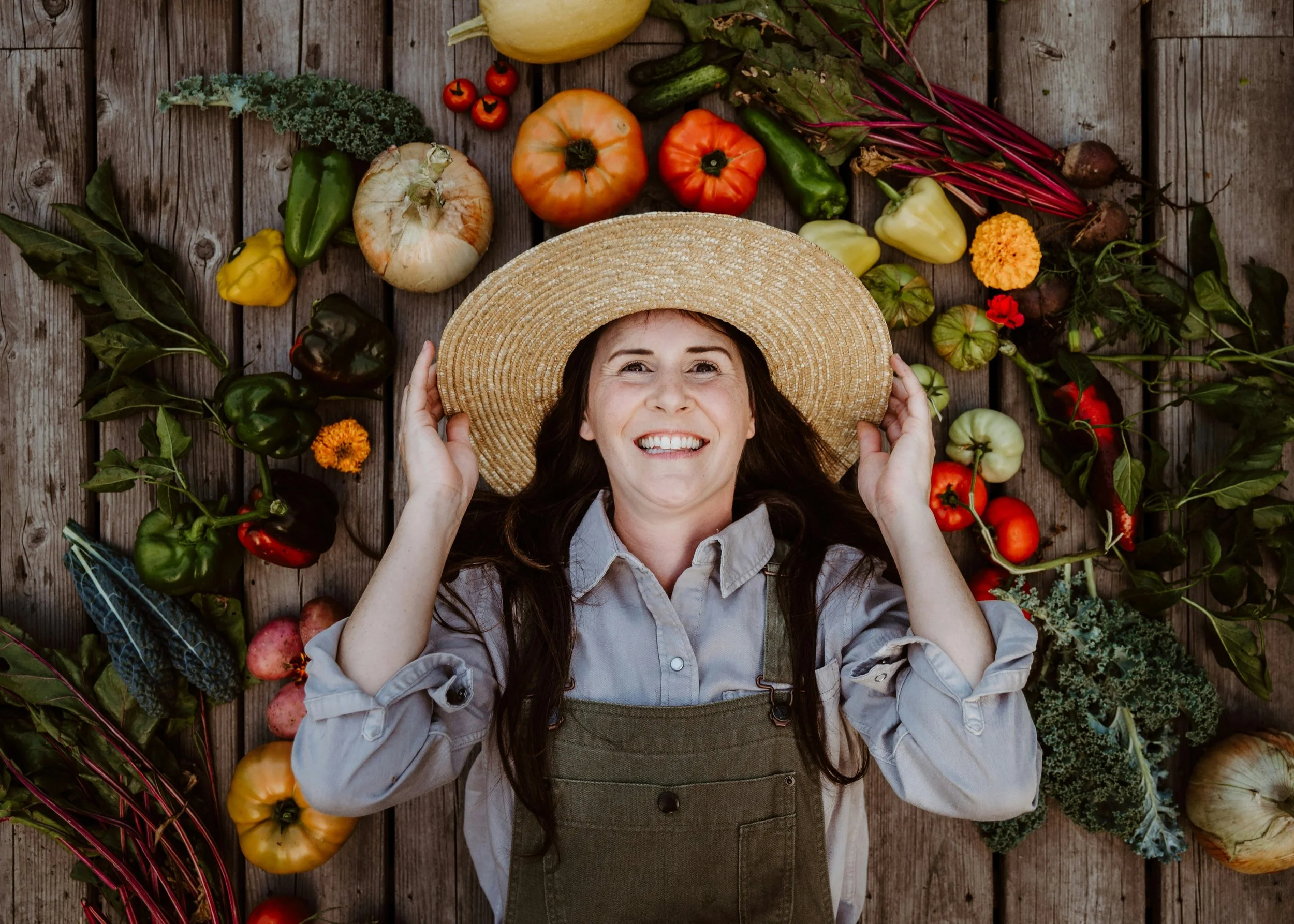 Een vrouw ligt op een houten vloer omringd door diverse groenten en fruit, terwijl ze een strohoed op haar hoofd houdt en glimlacht.