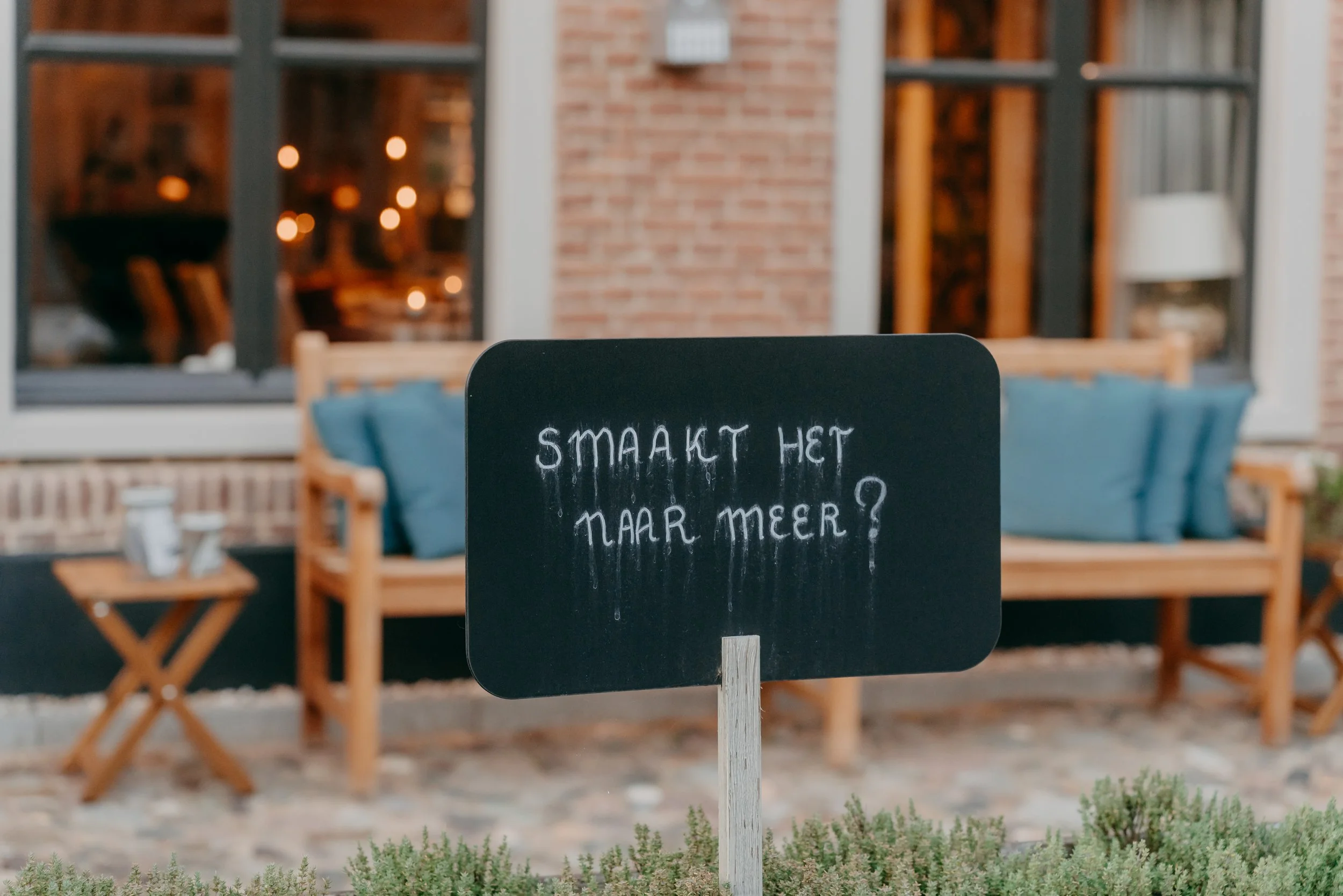 Een zwart bord met witte tekst dat vraagt 'Smaakt het naar meer?' voor een terras met blauwe kussens en een bank, buiten onder een raam met bruin bakstenen muur.