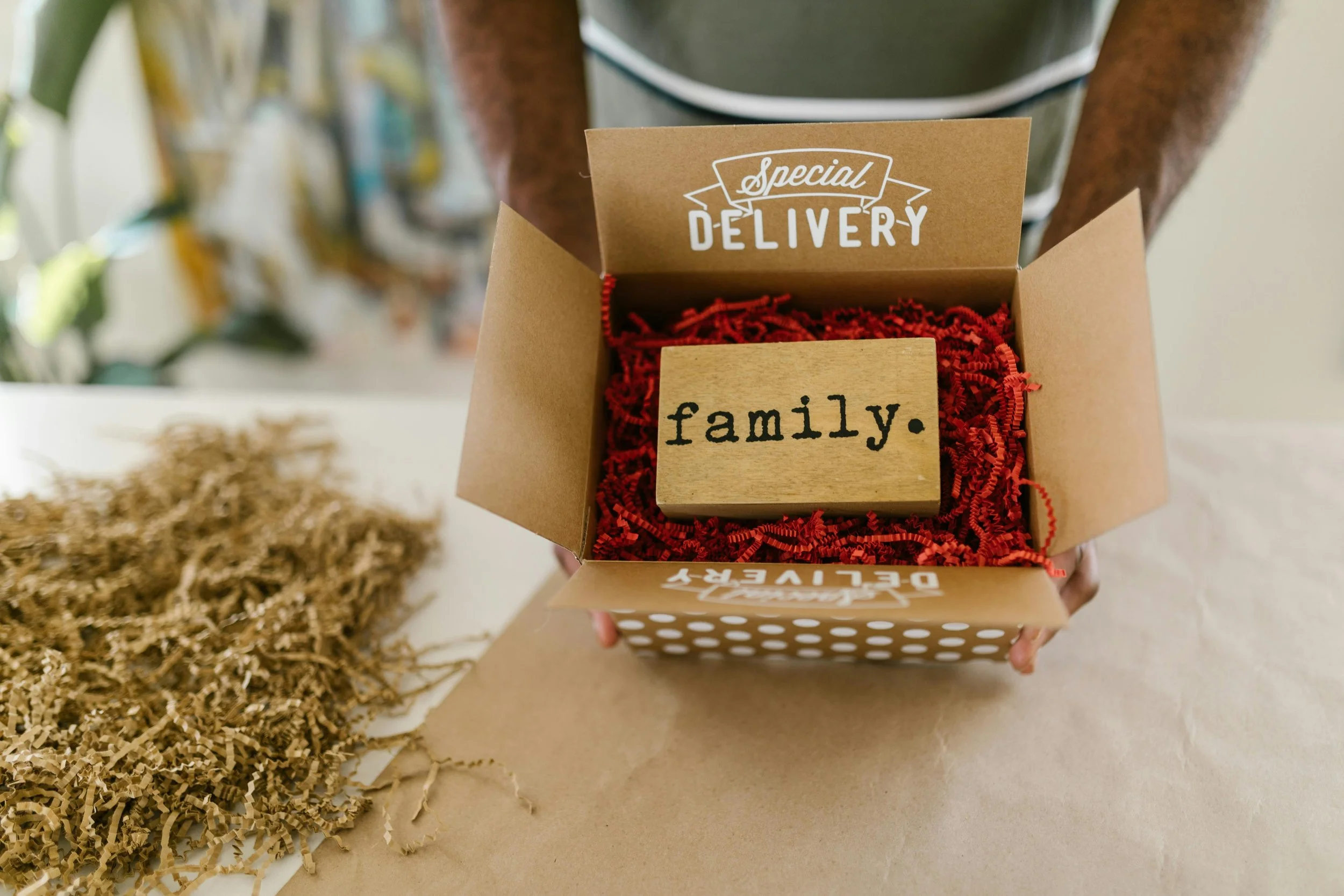 Persoon houdt een cadeauverpakking met het woord 'family' op een houten blok. Binnenin de doos ligt rood verpakkingsmateriaal en een sticker met 'Special Delivery' op de rand.
