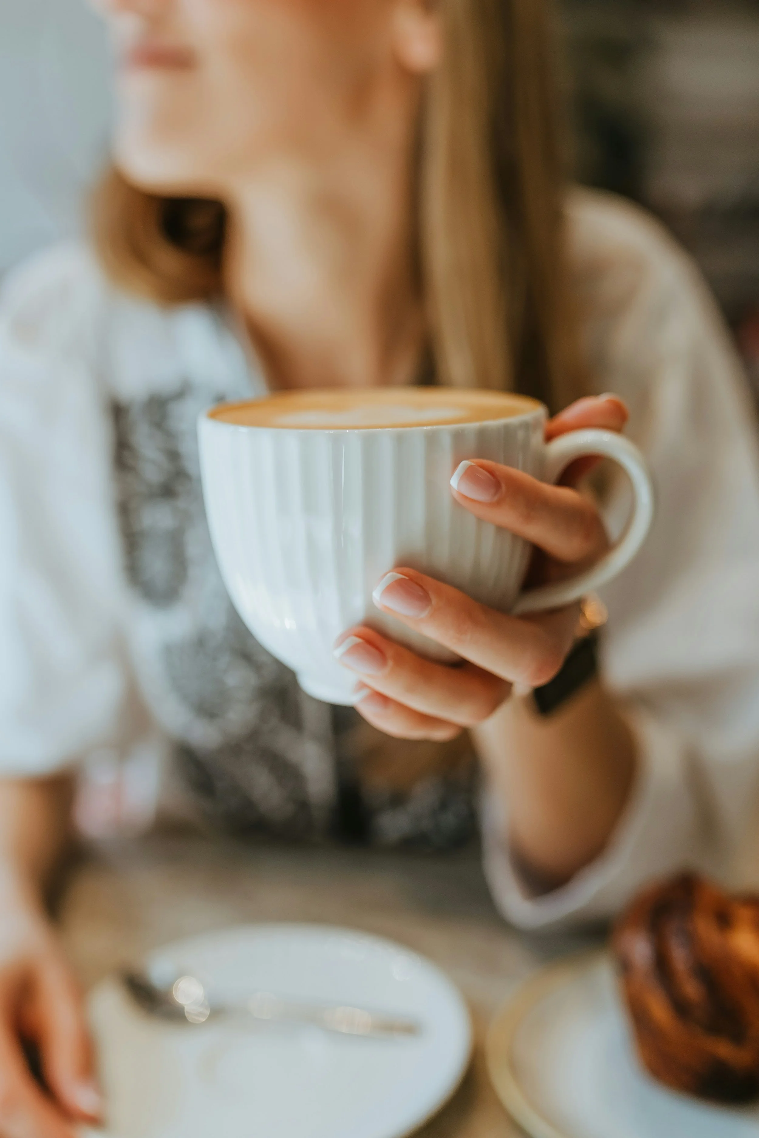 Een vrouw houdt een kop koffie vast met een warme drank op een tafel, omgeven door andere koppen en eten. Ze draagt een witte blouse met een zwarte detail en heeft lang, blond haar.