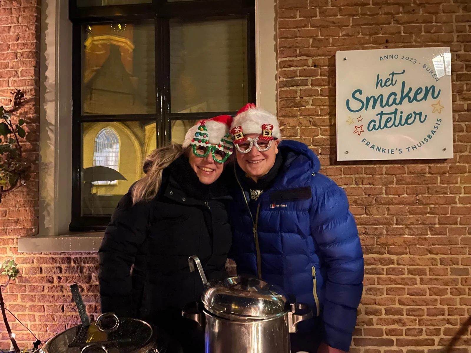 Twee mensen dragen kerstpetten en genialistische brillen, lachen, staan naast een grote pot in een buitenomgeving met een bakstenen muur en een raam op de achtergrond.