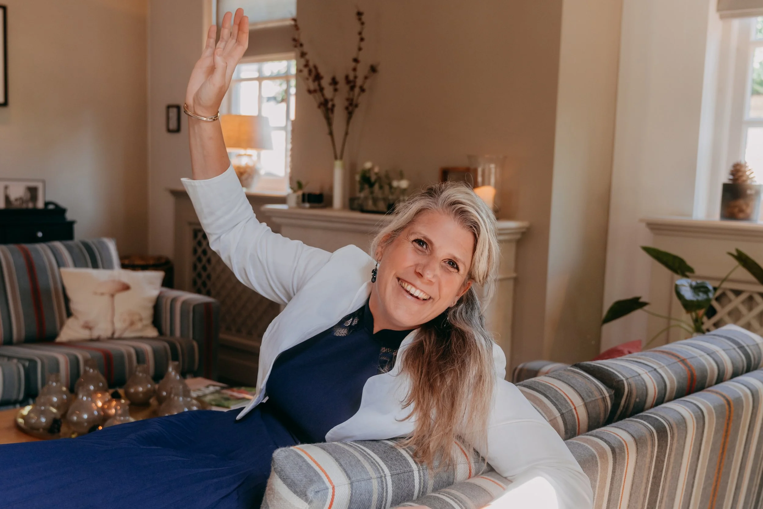 Vrolijke vrouw met lang blond haar zwaait vanuit een woonkamer met neutrale kleuren en planten.