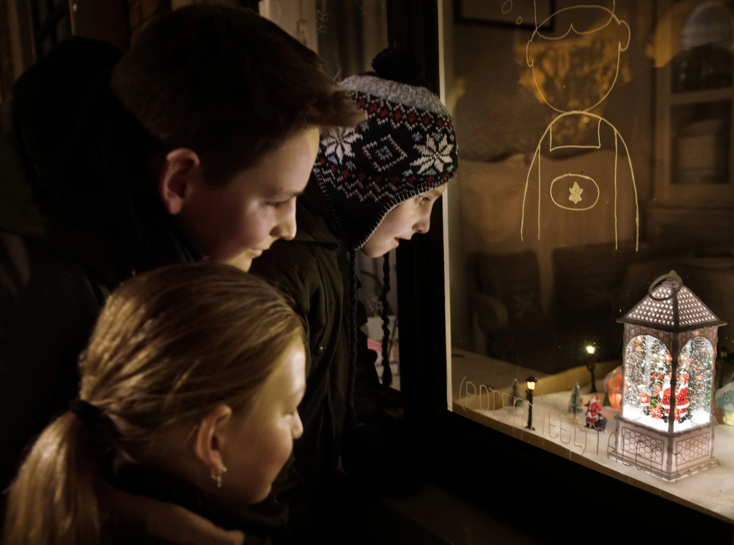 Drie kinderen kijken naar een kerststal door een raam met lichtjes en kerstdecoraties.