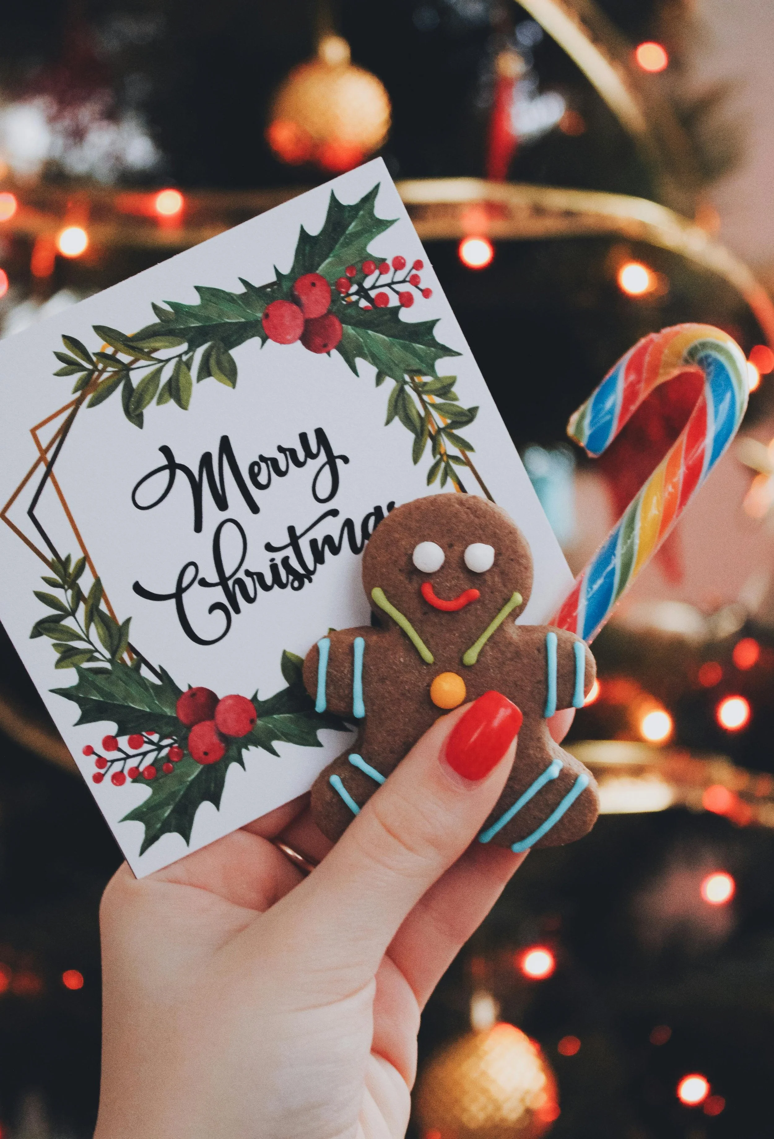 Een hand houdt een kerstkaart met de tekst 'Merry Christmas', versierd met een kersttak en besjes, samen met een gingerbread man koekje en een gekleurde candy cane op een kerstboom achtergrond.