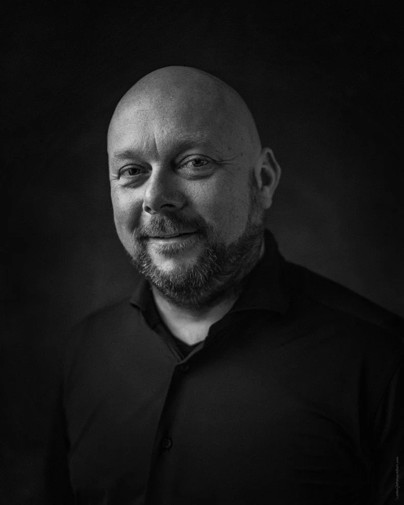 Zwart-wit portret van een baldere man met een baard, lachend en gekleed in een zwarte blouse, voor een donkere achtergrond.