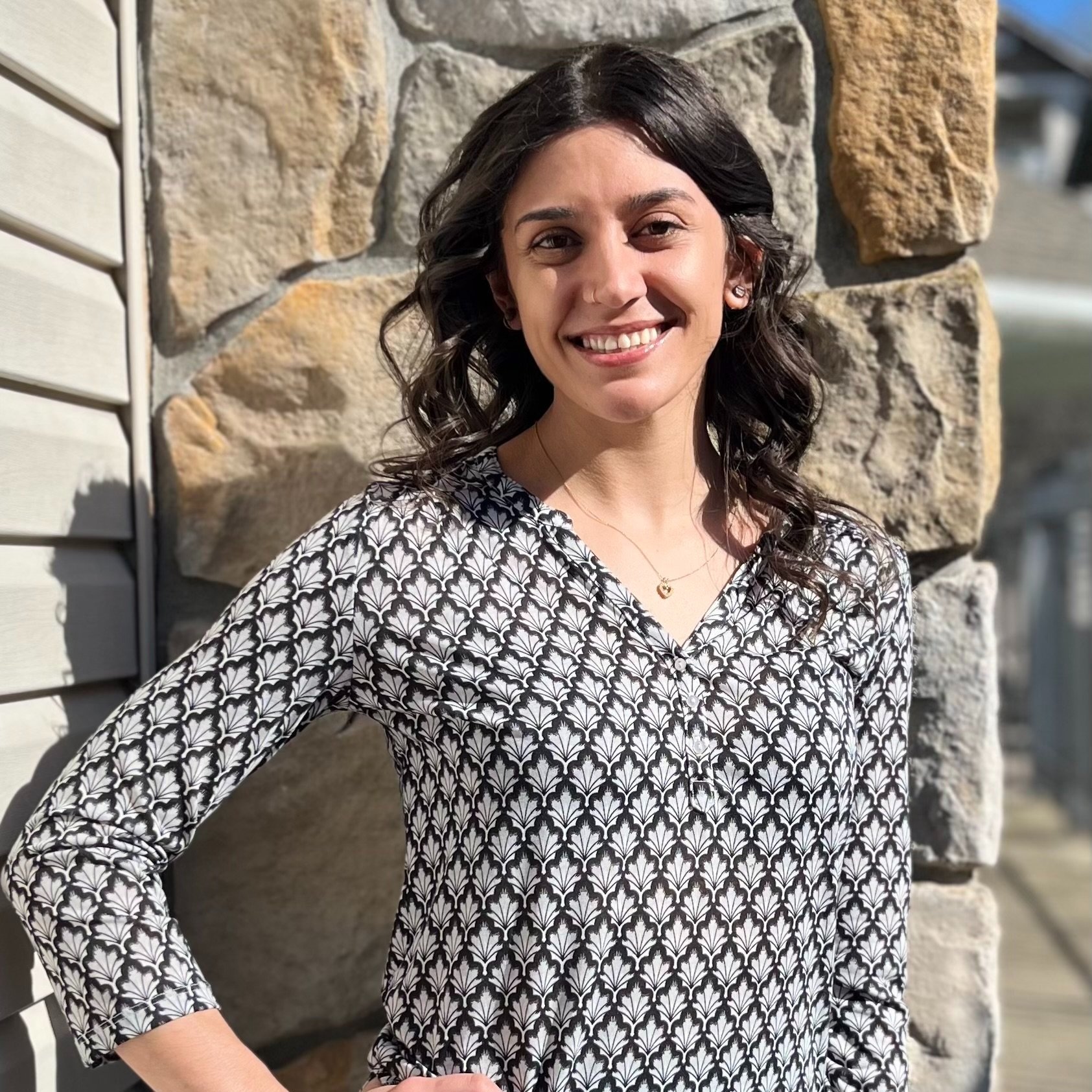 A woman with dark wavy hair smiling, wearing a black and white patterned blouse, standing outdoors against a stone wall.