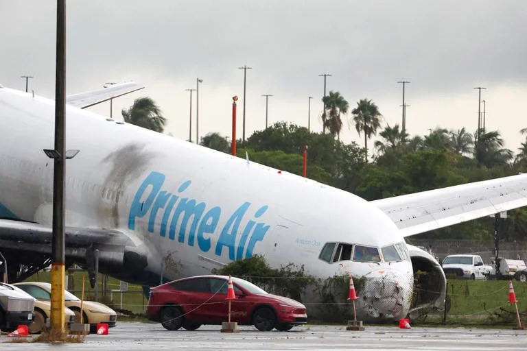 A large, damaged airplane with 'PrimeAir' branding on the fuselage, sitting on a tarmac near several cars and orange traffic cones, under overcast skies.