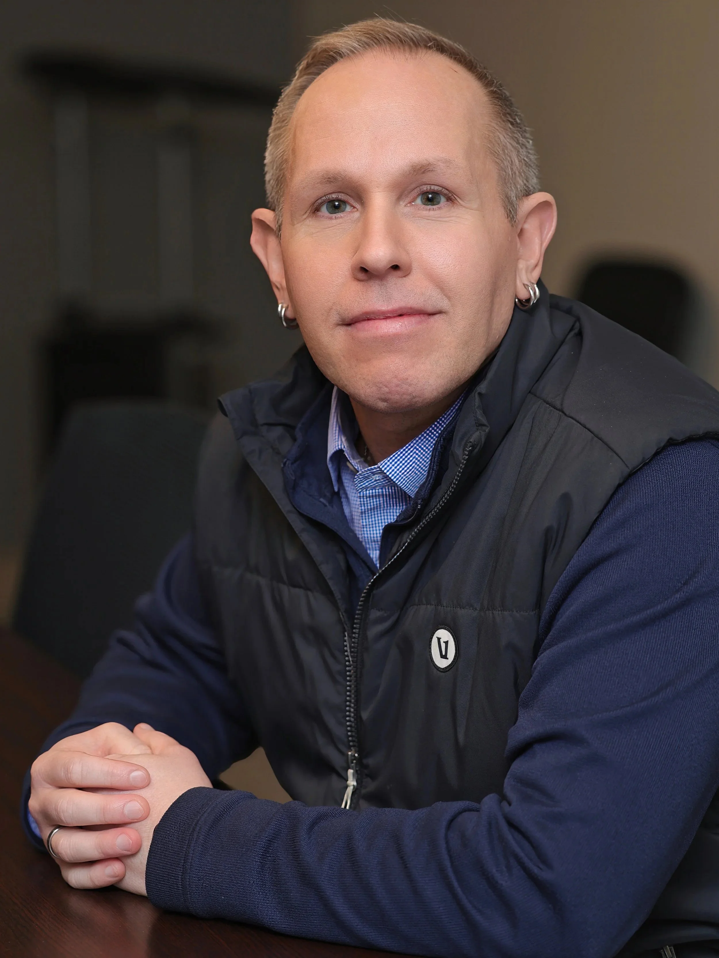 A man with short blond hair, pierced ears, and light skin, sitting at a table in a casual indoor setting. He is wearing a blue checkered shirt and a black vest. His hands are clasped together on the table, and he is looking directly at the camera with a slight smile.