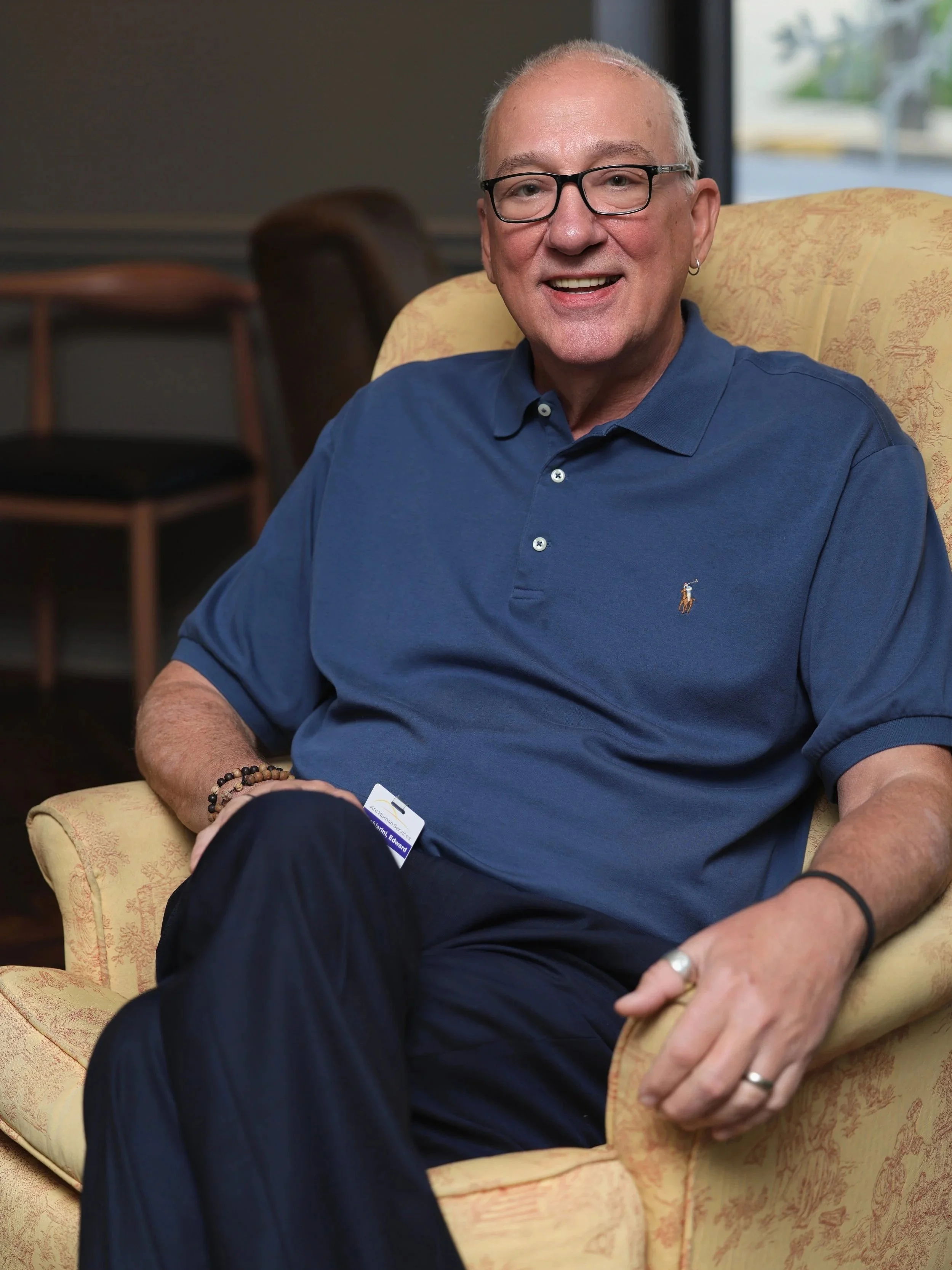 A smiling older man with glasses, a blue polo shirt, and rings on his finger, sitting comfortably in a yellow patterned armchair in a well-lit room.