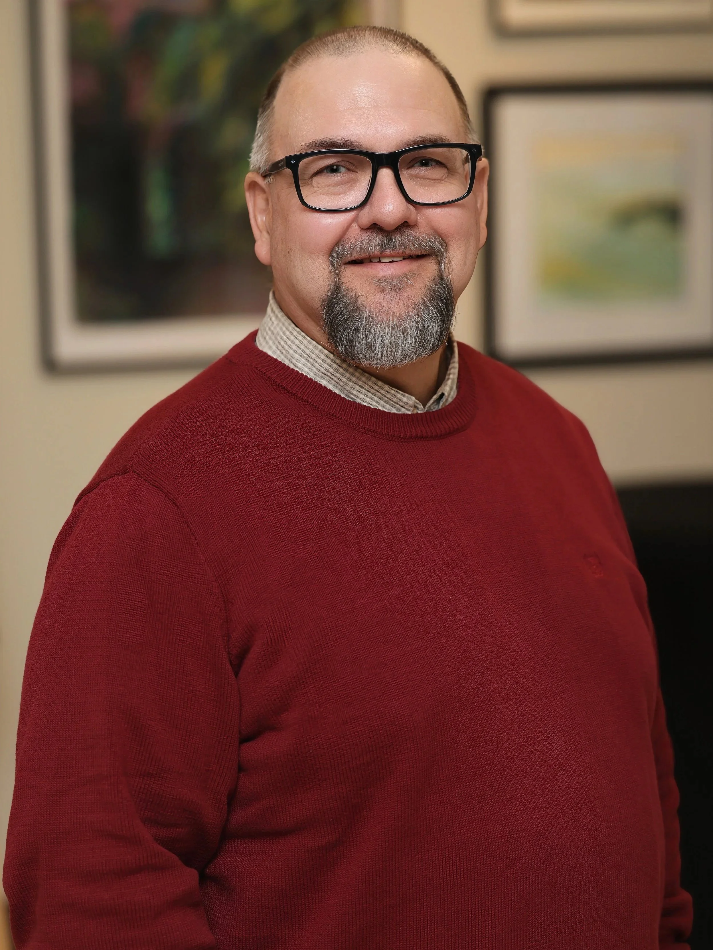 A middle-aged man with glasses, a beard, and a mustache standing indoors. He is wearing a red sweater over a collared shirt. There are framed pictures on the wall behind him.
