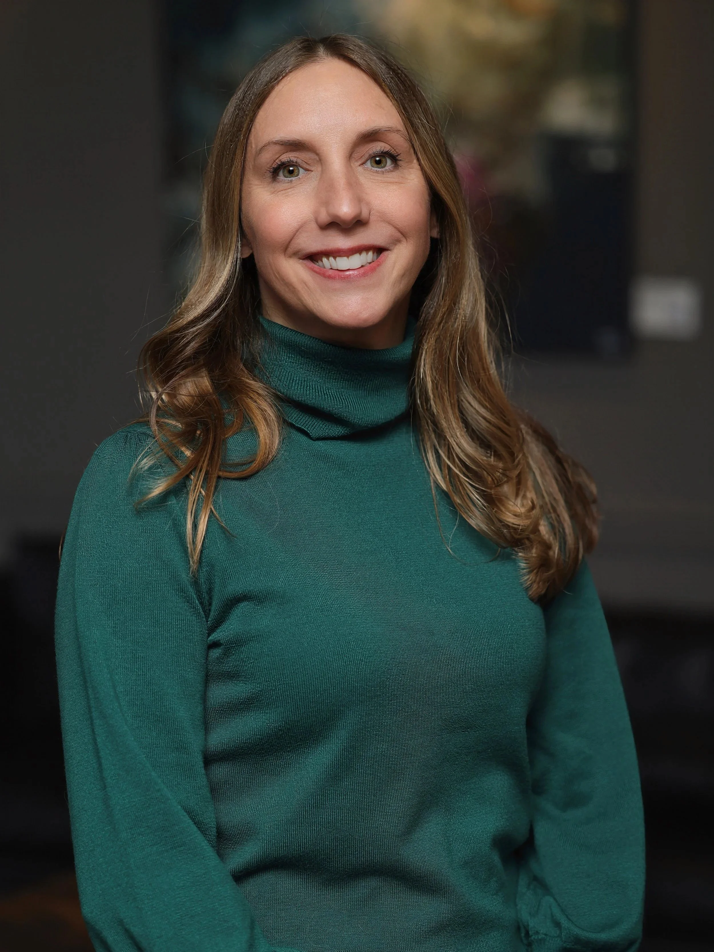A woman with long light brown hair wearing a teal turtleneck sweater, smiling with her teeth showing, standing in an indoor setting with a blurred background.
