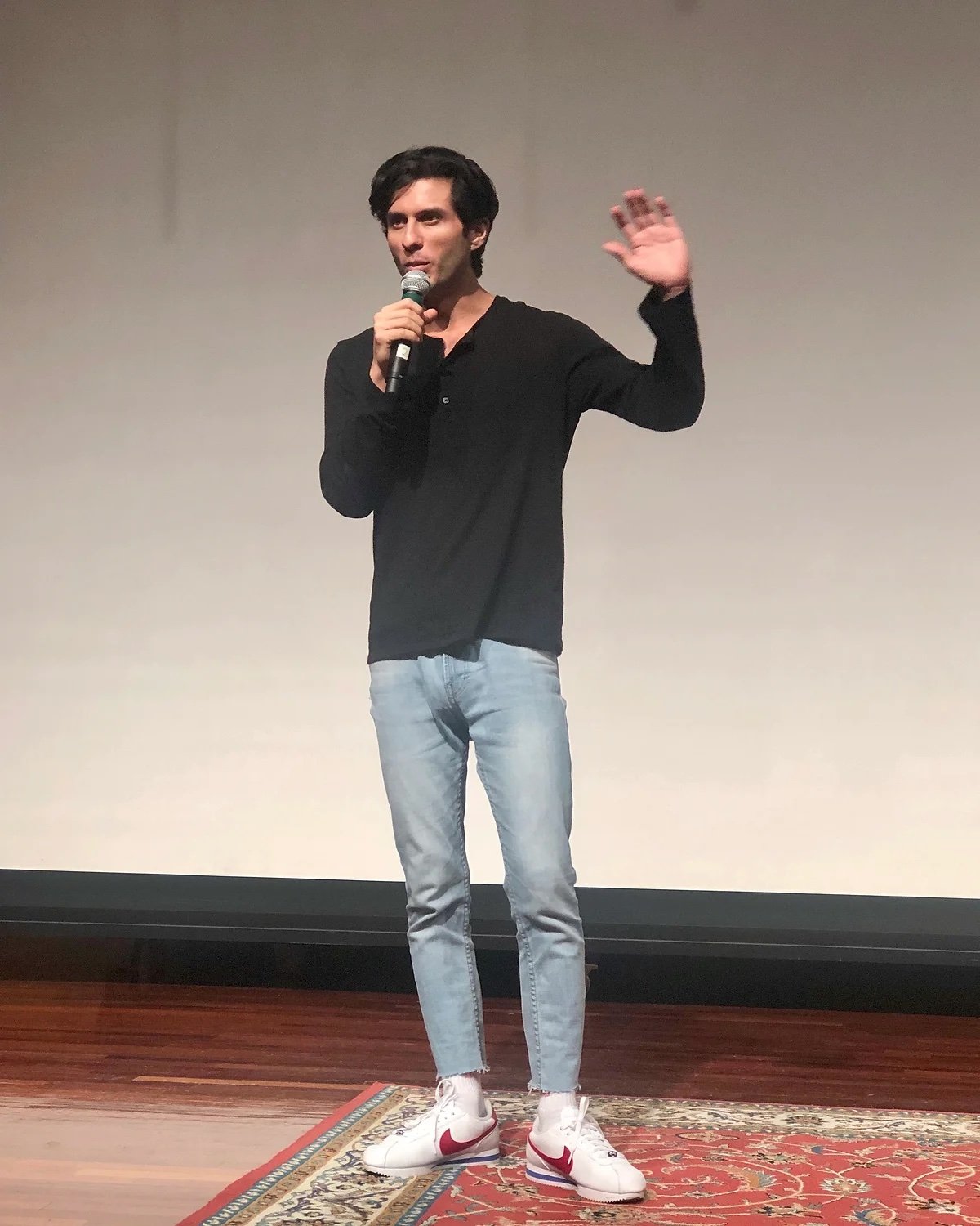 A young man with dark hair and light skin standing on a stage, speaking into a microphone and waving. He's wearing a black long-sleeve shirt, light blue jeans, and white Nike sneakers with red swooshes. The background is plain and the stage has a woo