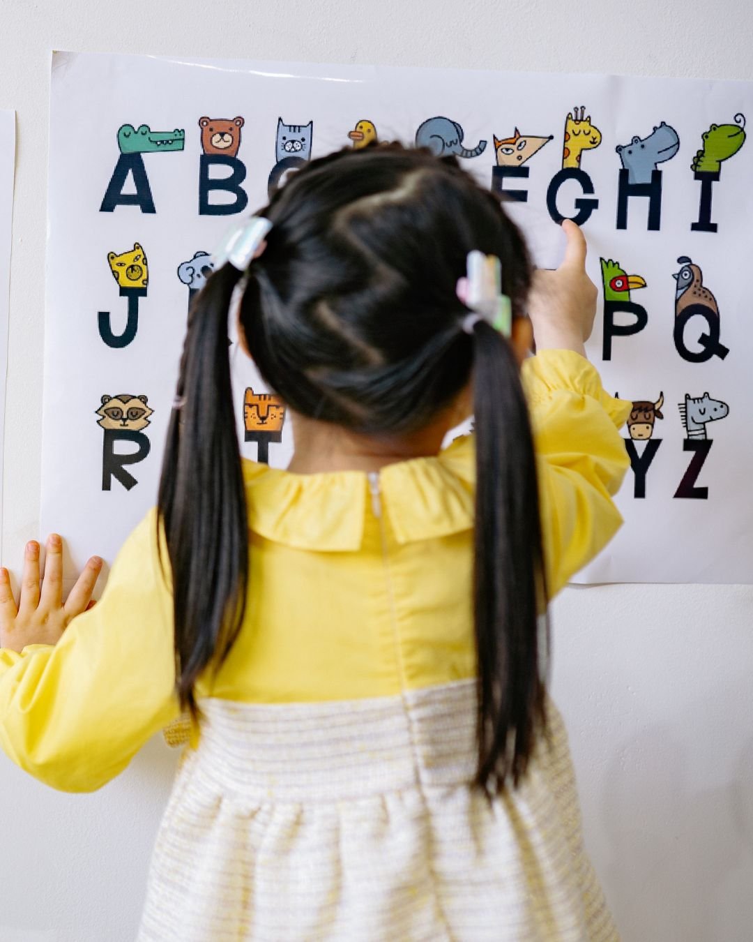 Child in yellow jacket with pigtails pointing at an educational poster of the alphabet with animal illustrations.