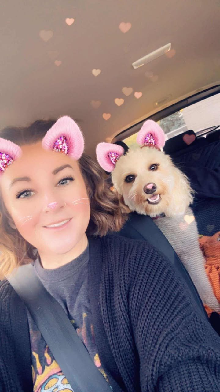A woman and a goldendoodle taking a selfie in a car, both wearing pink, glittery animal ear filters and having fun.