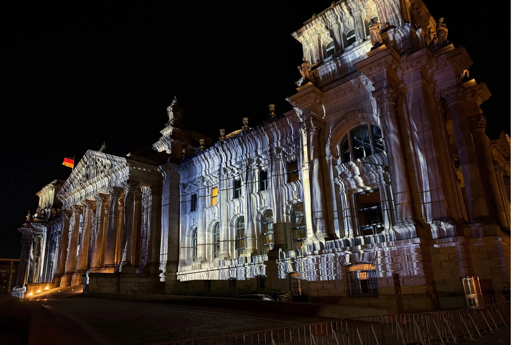 Front exterior of the Reichstag building in Berlin at night, with dramatic light projections illuminating the stone facade and classical columns.