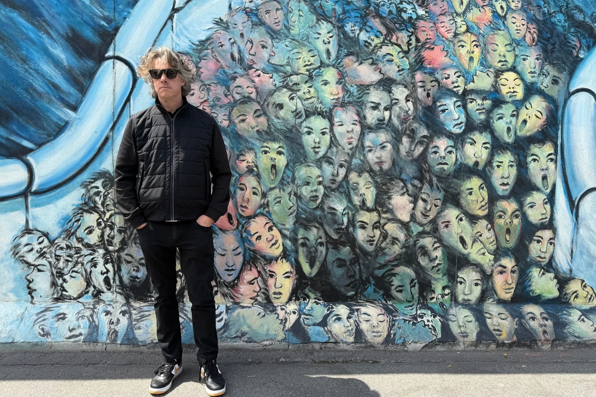 Jay standing by the It Happened in November mural at the East Side Gallery, surrounded by a multitude of painted faces.