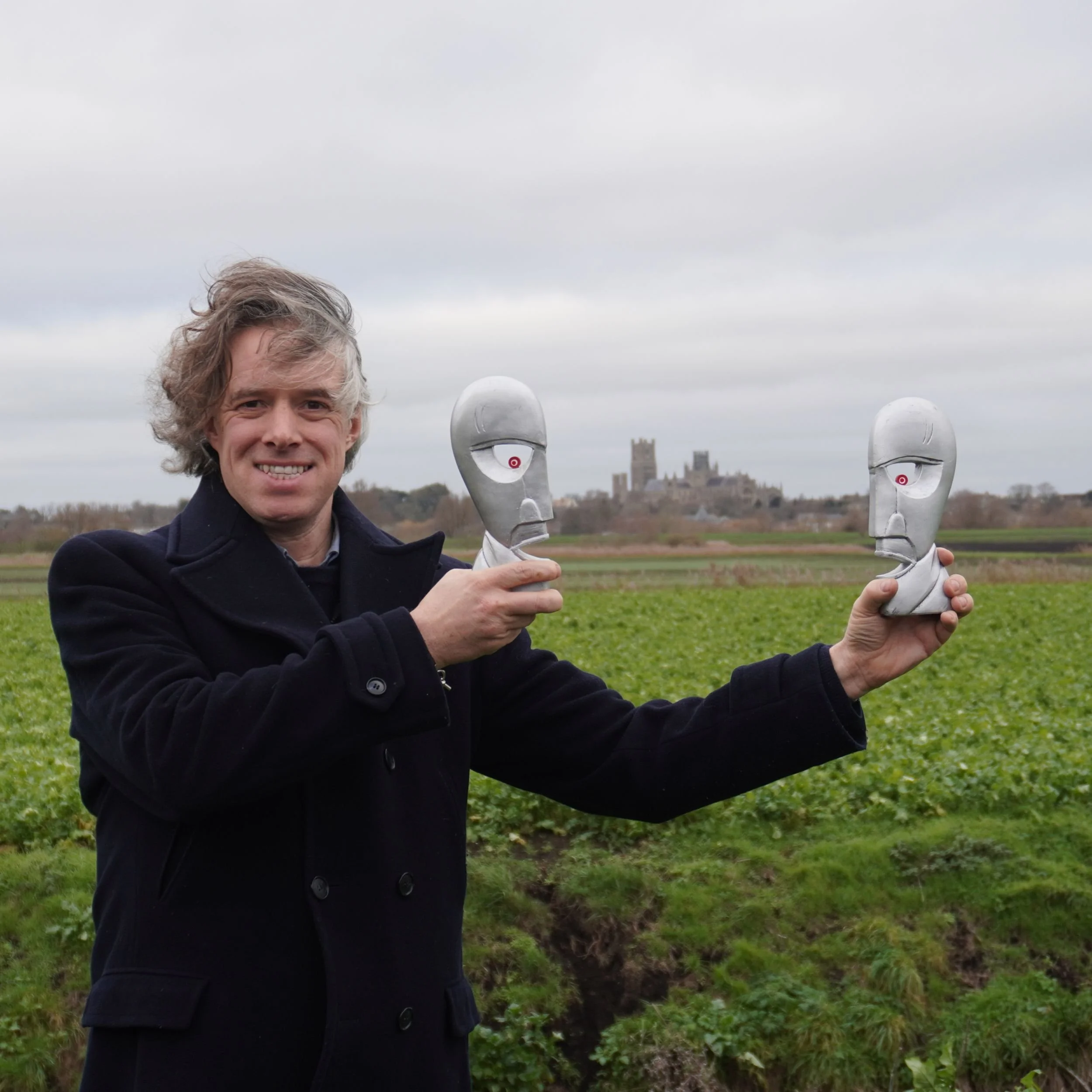 Replica Division Bell heads held apart in fenland fields outside Ely, showing the sightline towards Ely Cathedral