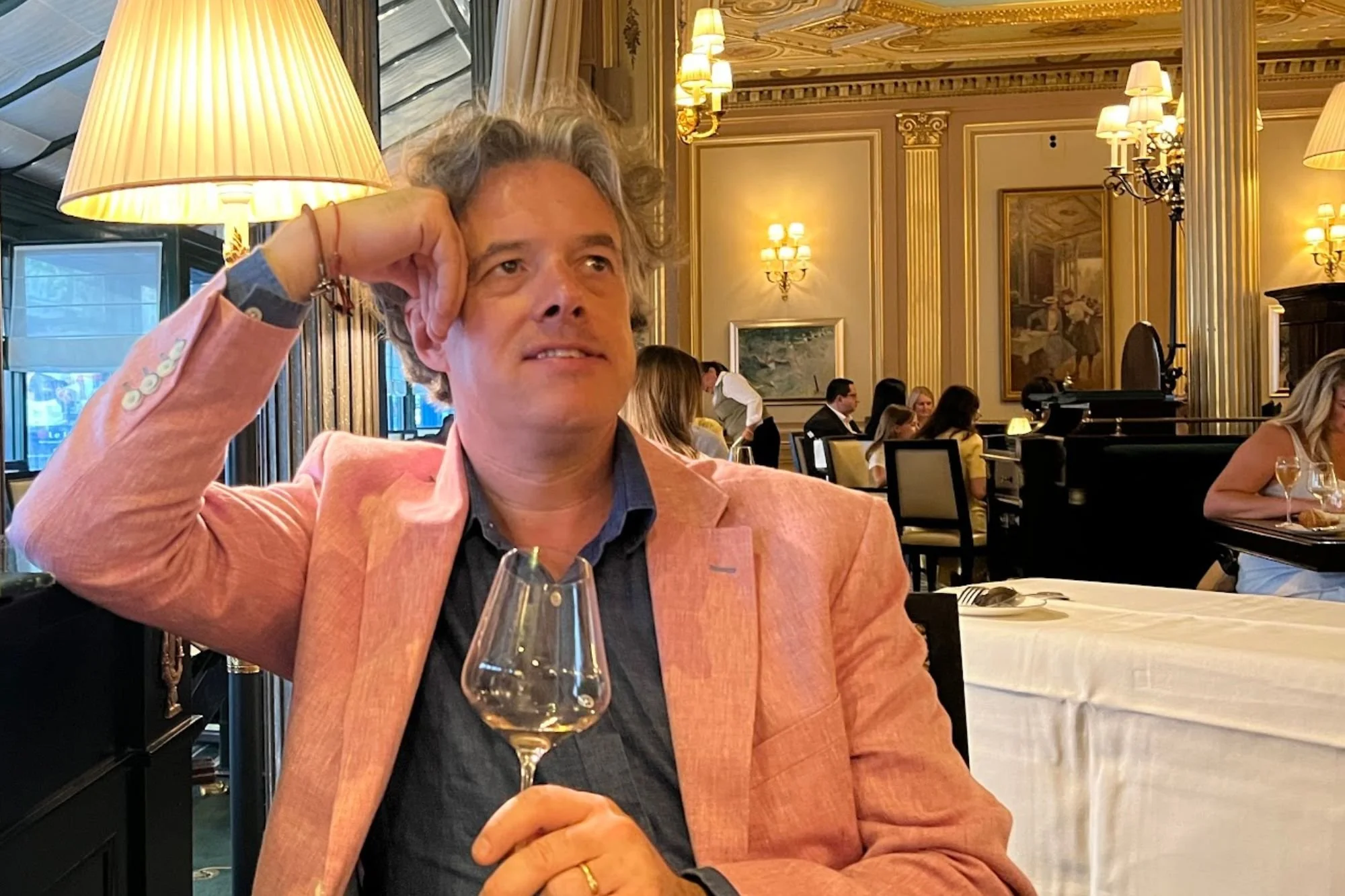 Jay in a pink linen blazer holding a glass of wine at a table inside Café de la Paix Paris