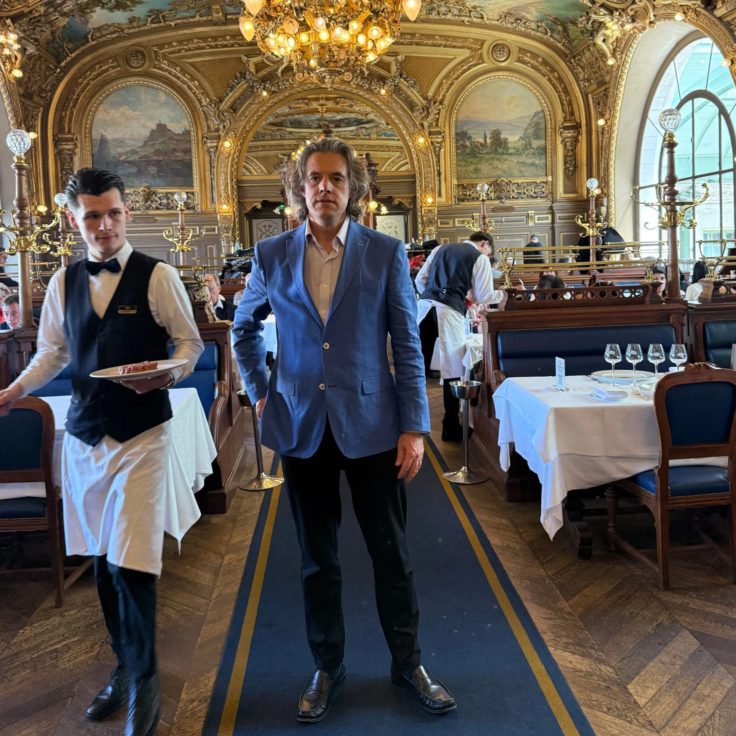 Guest wearing a blue jacket dining at Le Train Bleu in Paris, showing the smart but relaxed dress code inside the historic Gare de Lyon restaurant, with waitstaff and ornate Belle Époque interior visible.