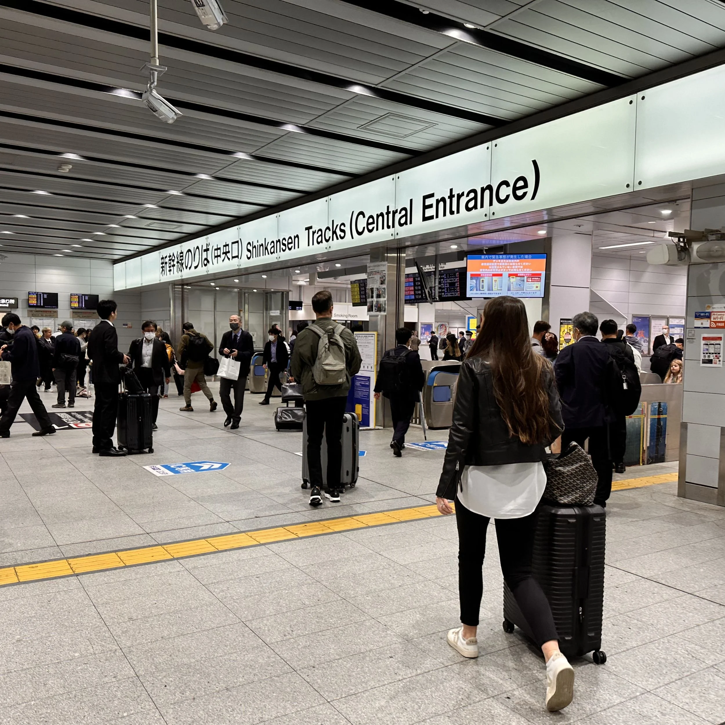 Travellers with suitcases heading towards the Shinkansen tracks at Shin-Osaka Station beneath a large bilingual station sign.