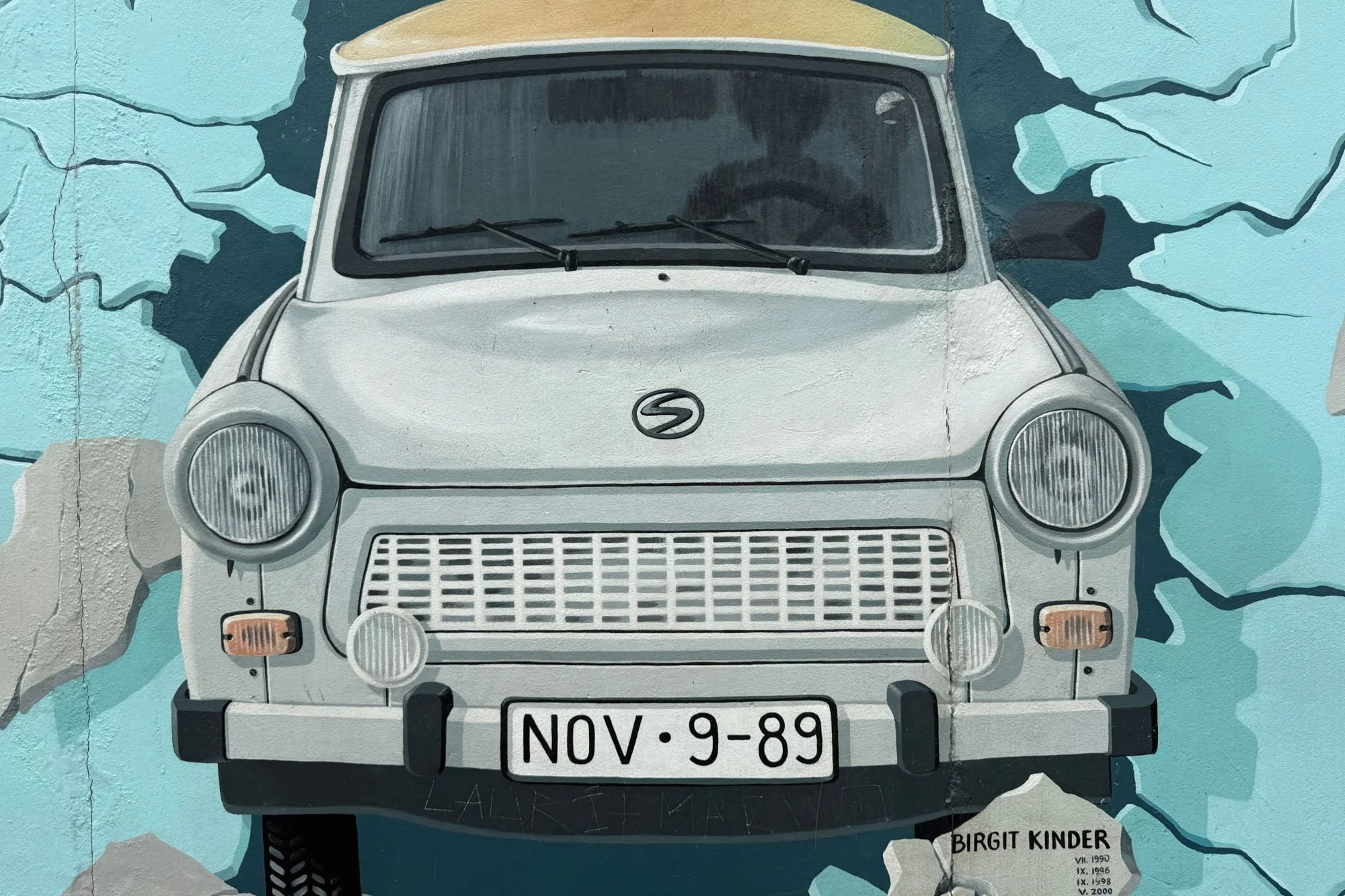 A close-up view of the Berlin Wall's Test the Rest mural by Birgit Kinder, featuring a grey Trabant car bursting through the concrete and a "NOV-9-89" licence plate.