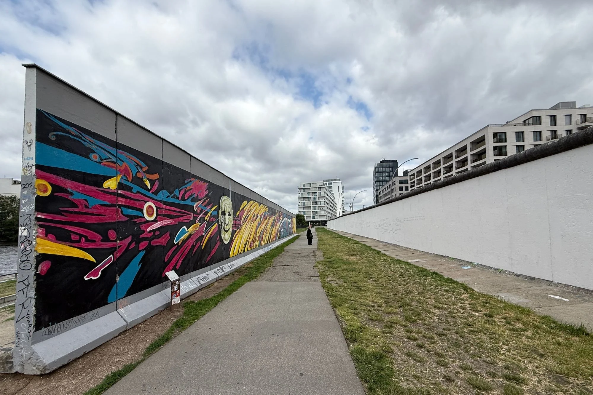A view looking between the outer painted panels of the East Side Gallery and the secondary concrete barrier of the original Berlin Wall, stepping into the grassy expanse of the former death strip.