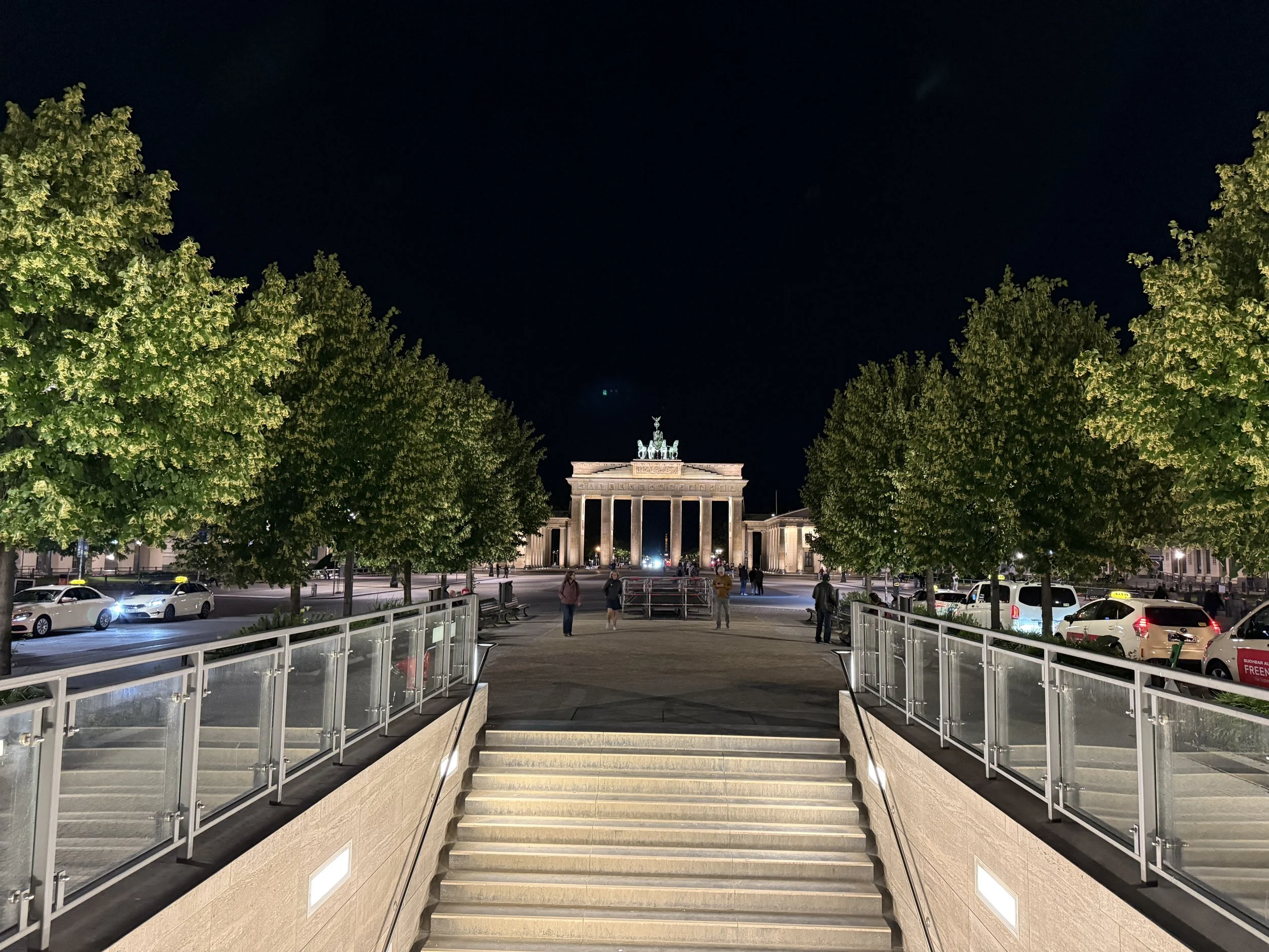 Wide stone entrance steps of Brandenburger Tor U-Bahn station framed by trees looking towards the illuminated Brandenburg Gate at night