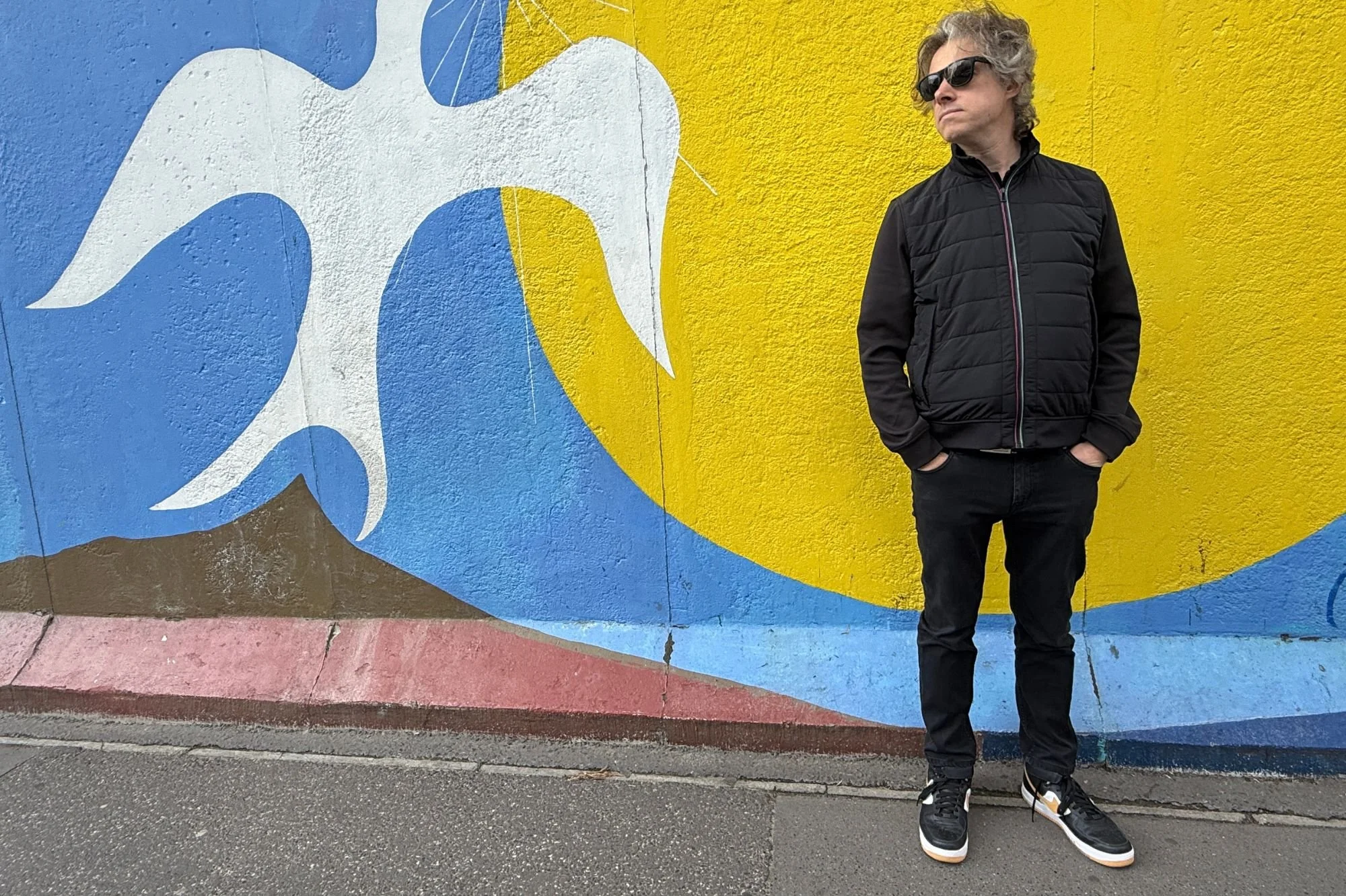 Jay standing beside The Dawn of Peace mural at the East Side Gallery, featuring a bold white bird against a yellow sun and blue sky.