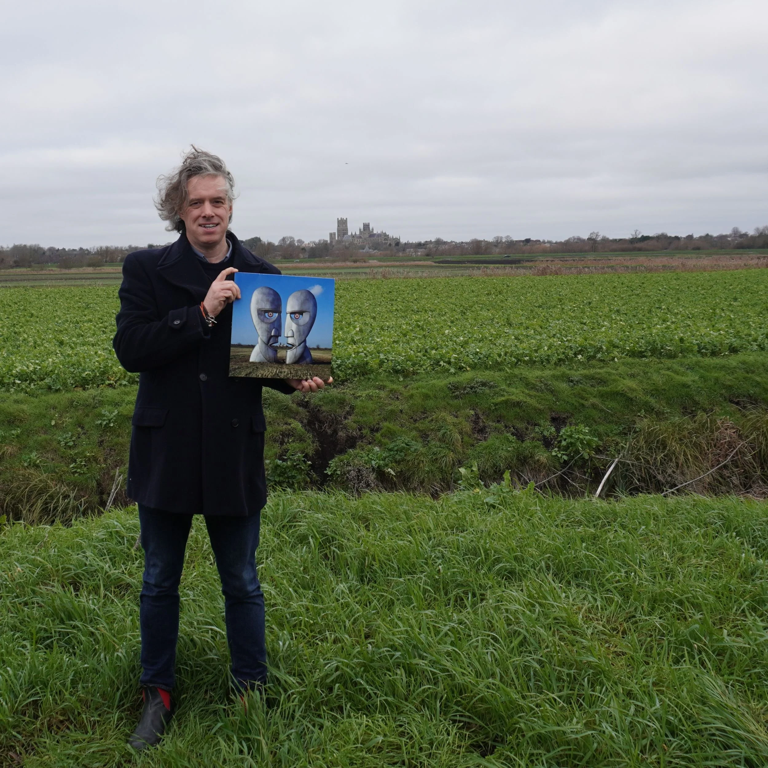 Jamie Riddell holding The Division Bell album in the Cambridgeshire Fens with Ely Cathedral visible in the distance