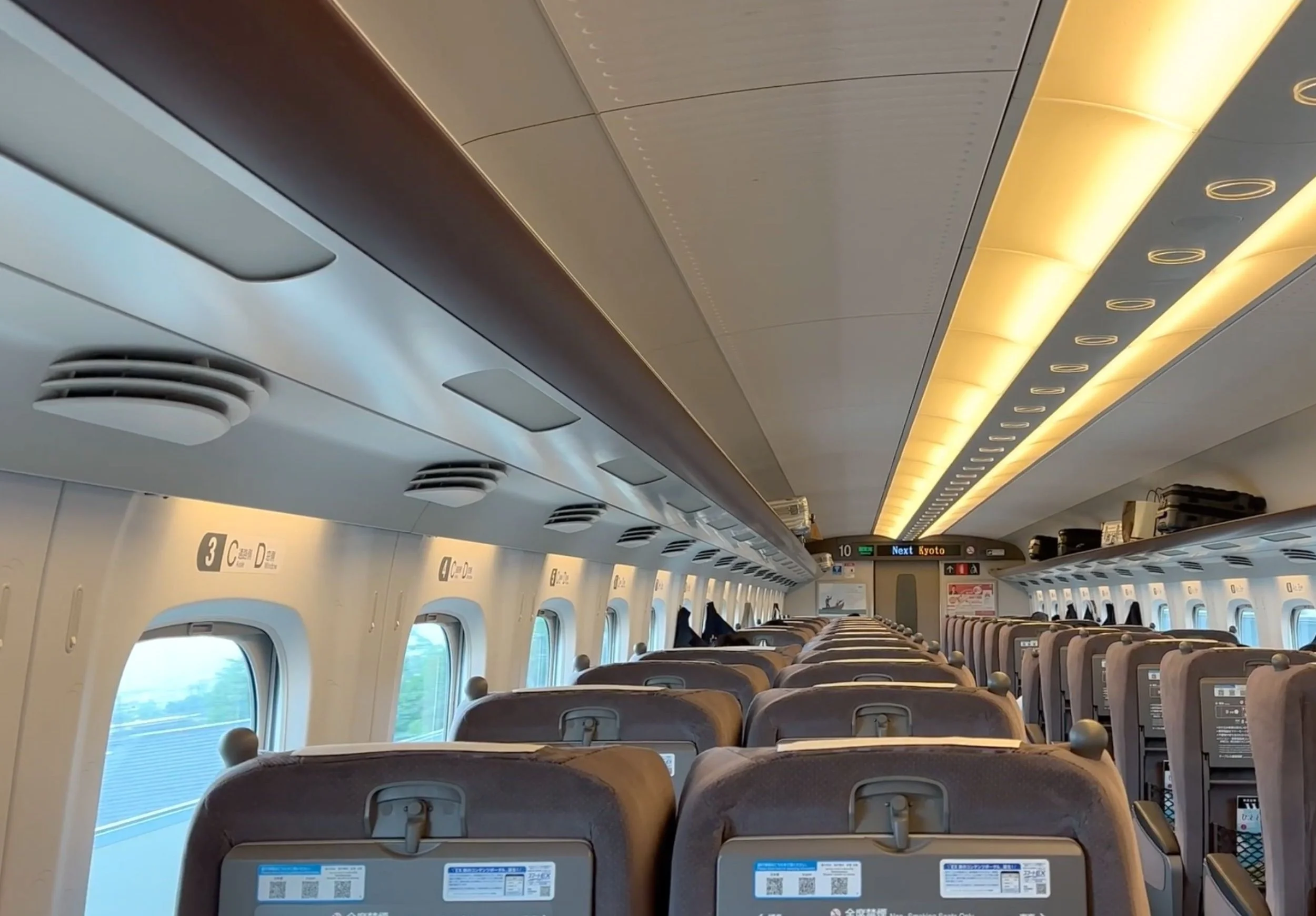 Interior of a Shinkansen carriage with rows of seats, overhead luggage racks and a digital sign showing the next stop as Kyoto.