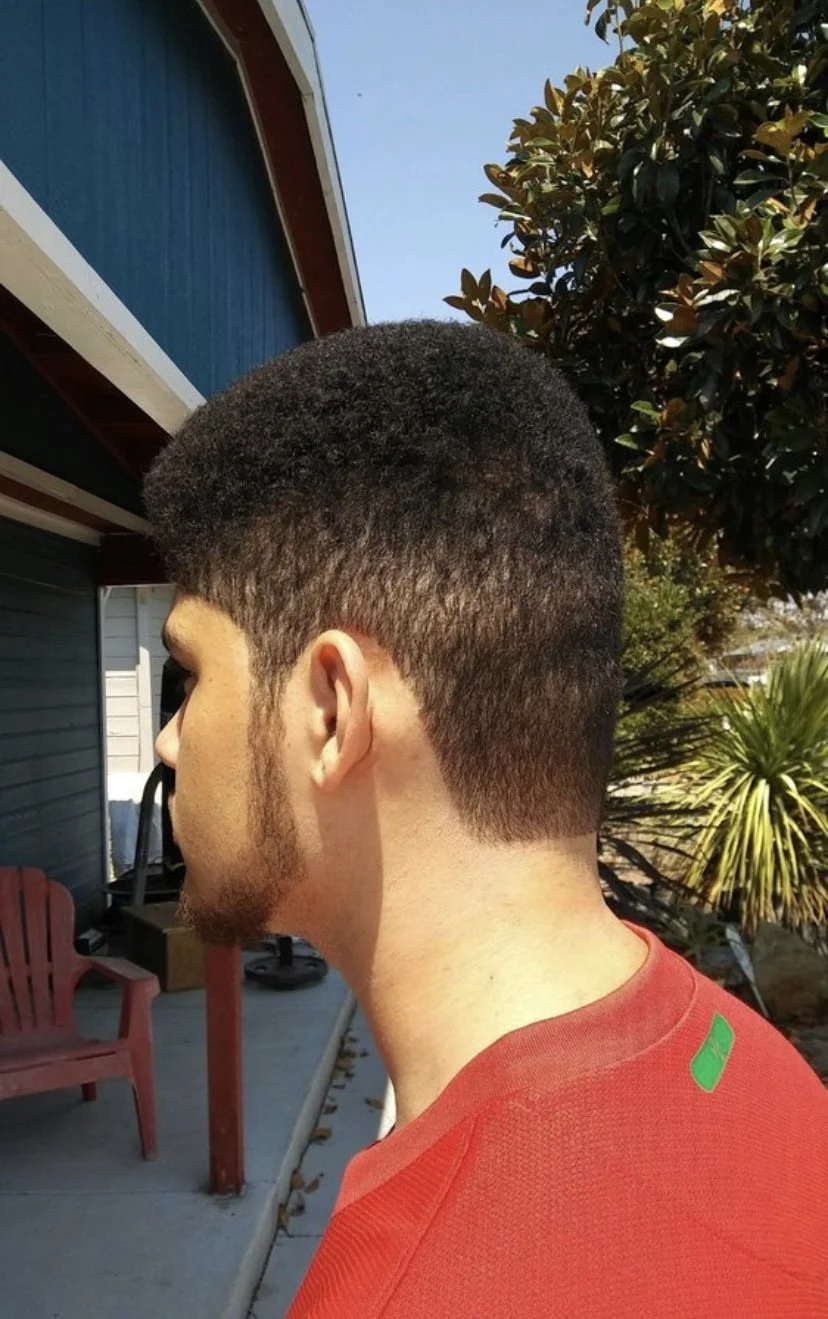 Side profile of a young man with a fade haircut wearing a red shirt, standing outdoors near plants and a house with blue siding.