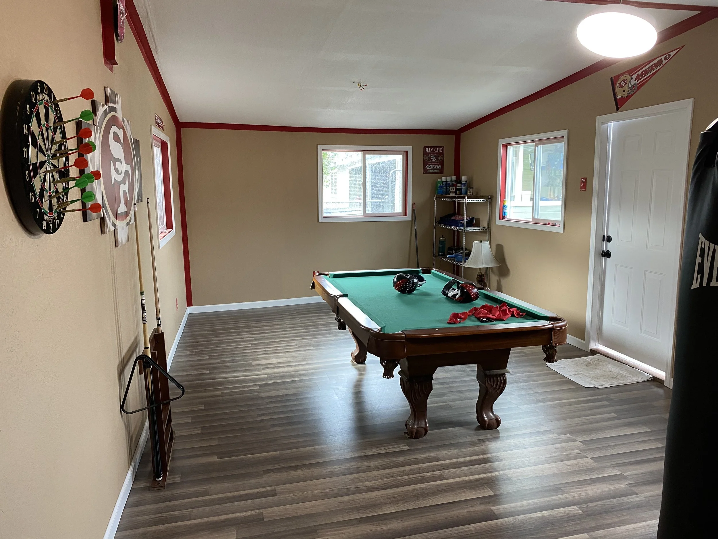 A recreational room with a pool table, dartboard, and sports memorabilia on the walls. Two windows, a shelving unit, and a lamp are visible, with a door and a mat on the floor.