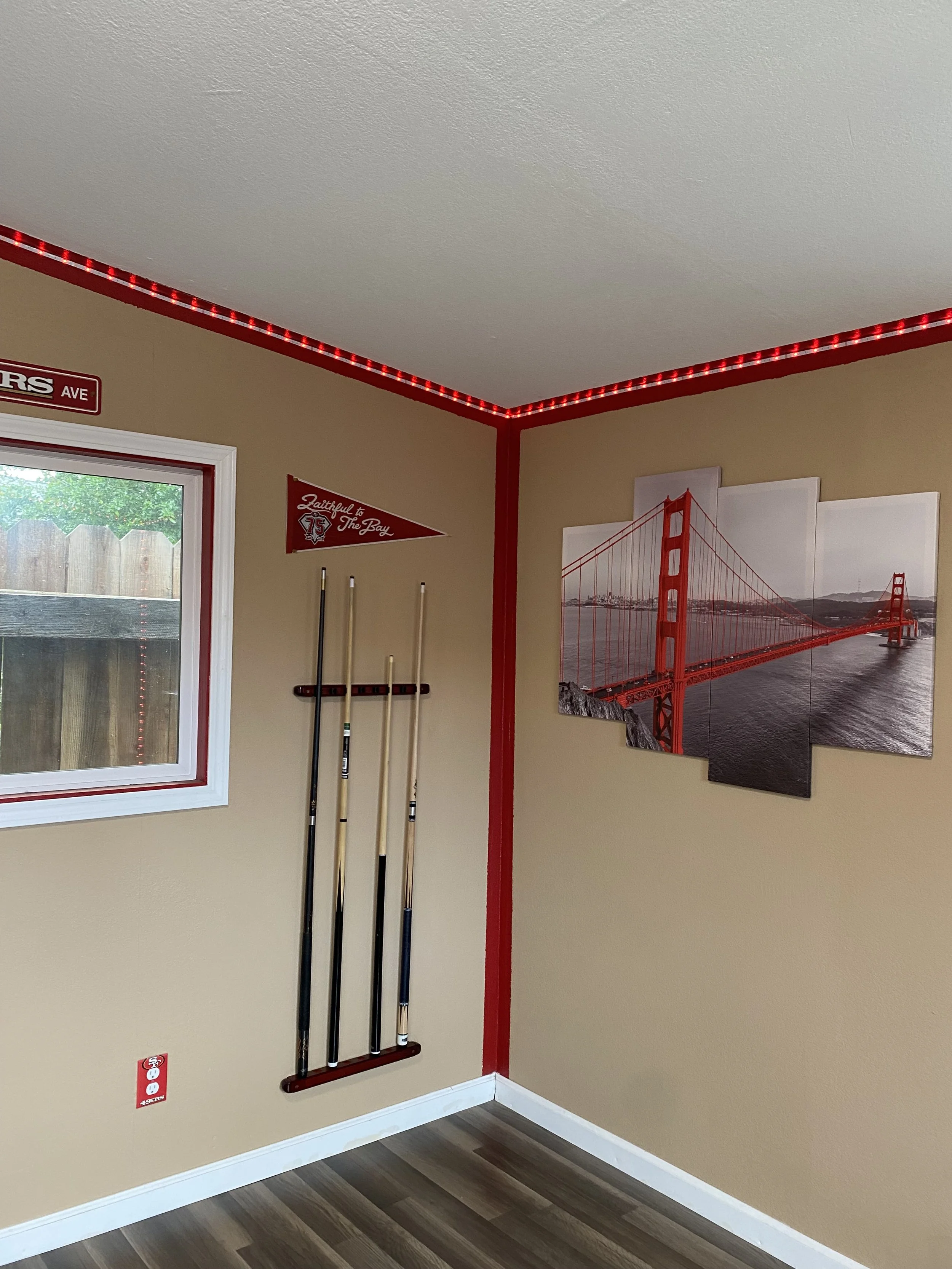Corner of a room with a window, a red string light or LED strip along the ceiling corner, a small red pennant with white text, a set of golf clubs mounted on the wall, and a multi-panel picture of the Golden Gate Bridge.
