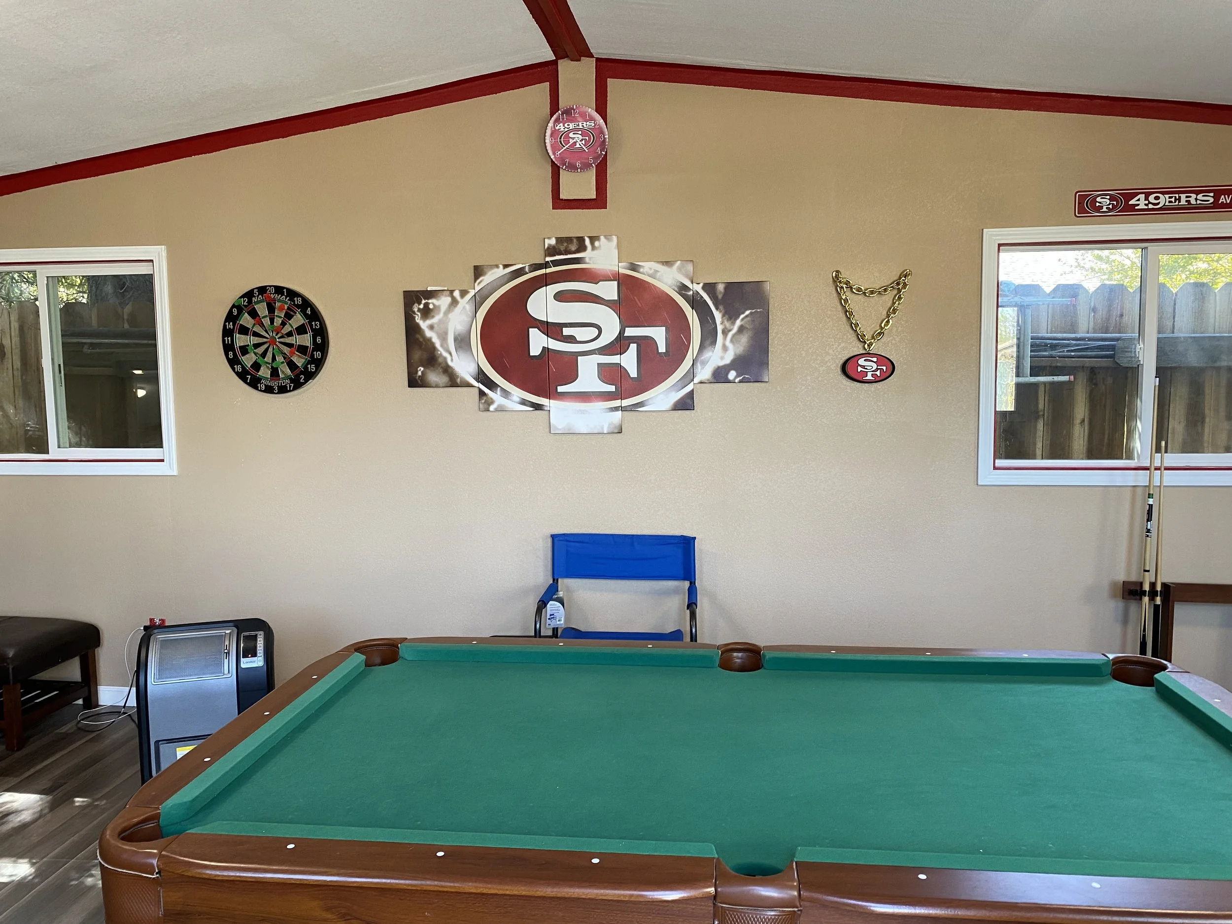 Room with a pool table, wall decorations including the San Francisco 49ers logo, dartboard, window, a blue chair, and a heater.